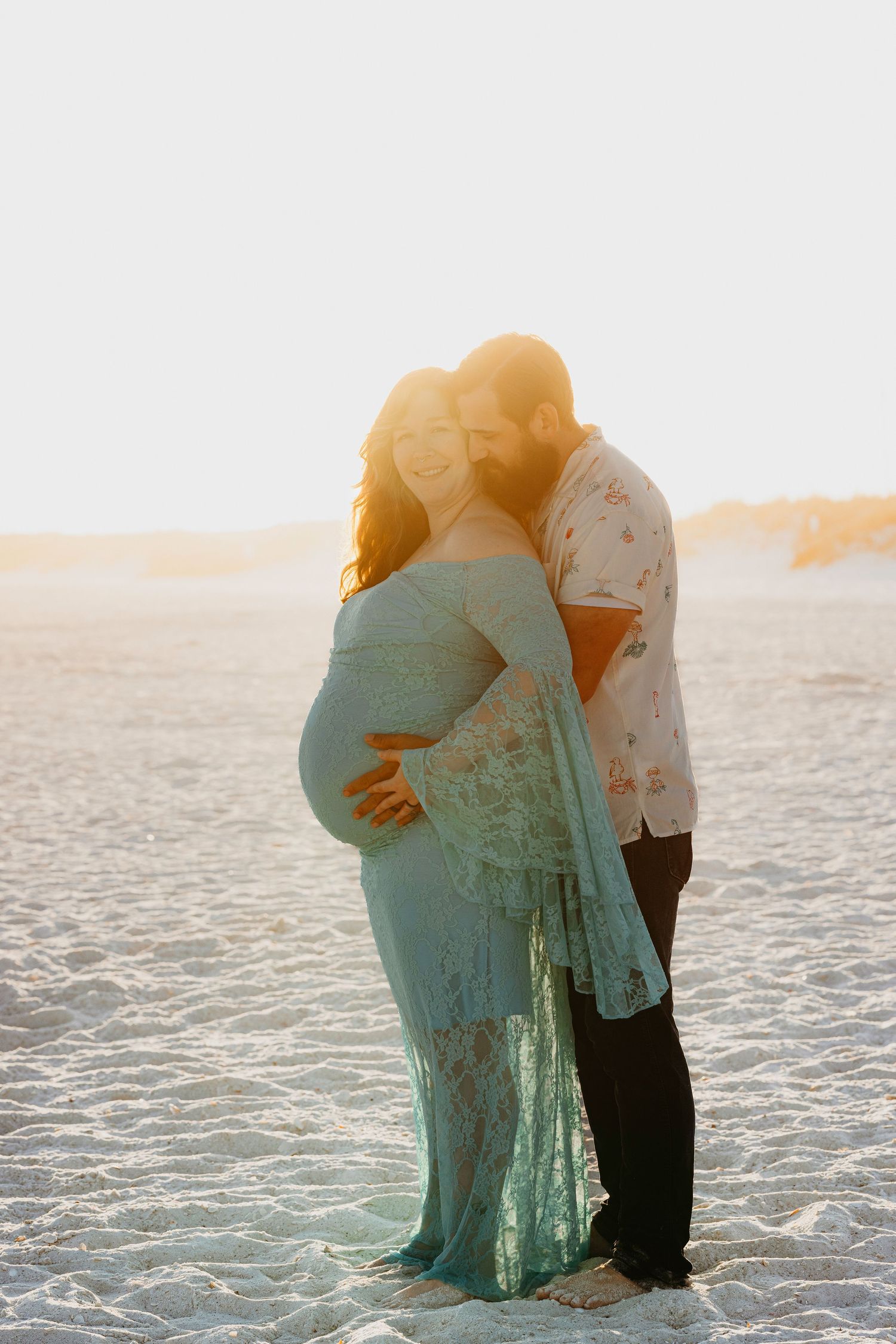 Glowing sunset maternity photo of couple embracing on beach, woman in teal lace gown.