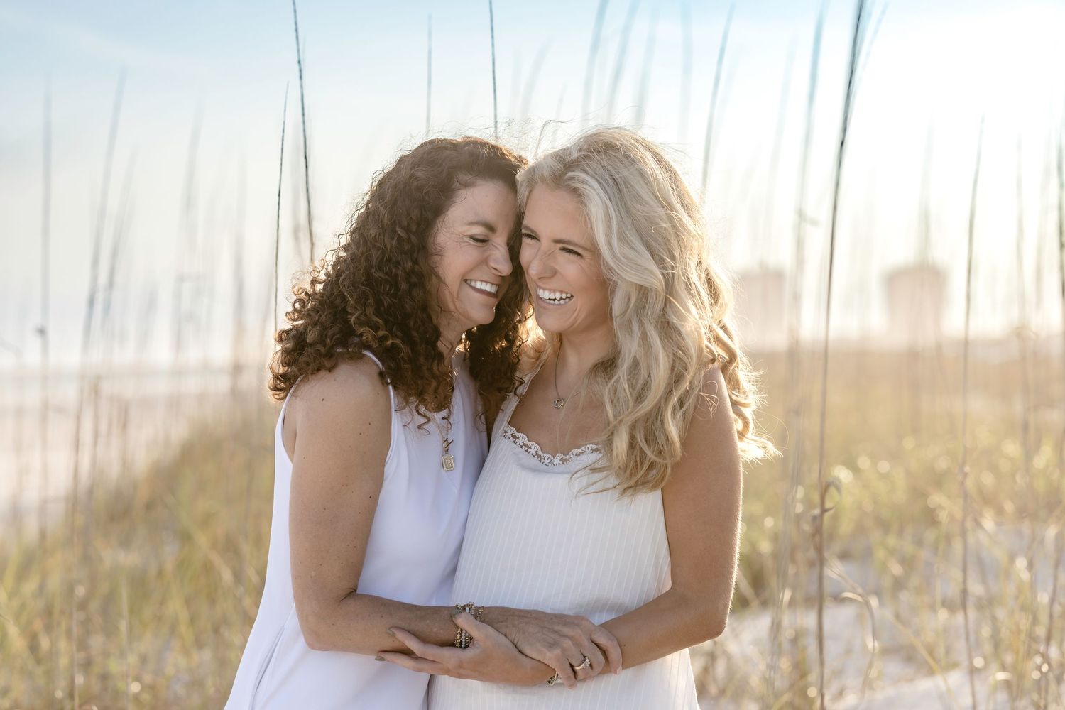Two women in white dresses laughing and embracing on a sunny beach with tall grass in the background.