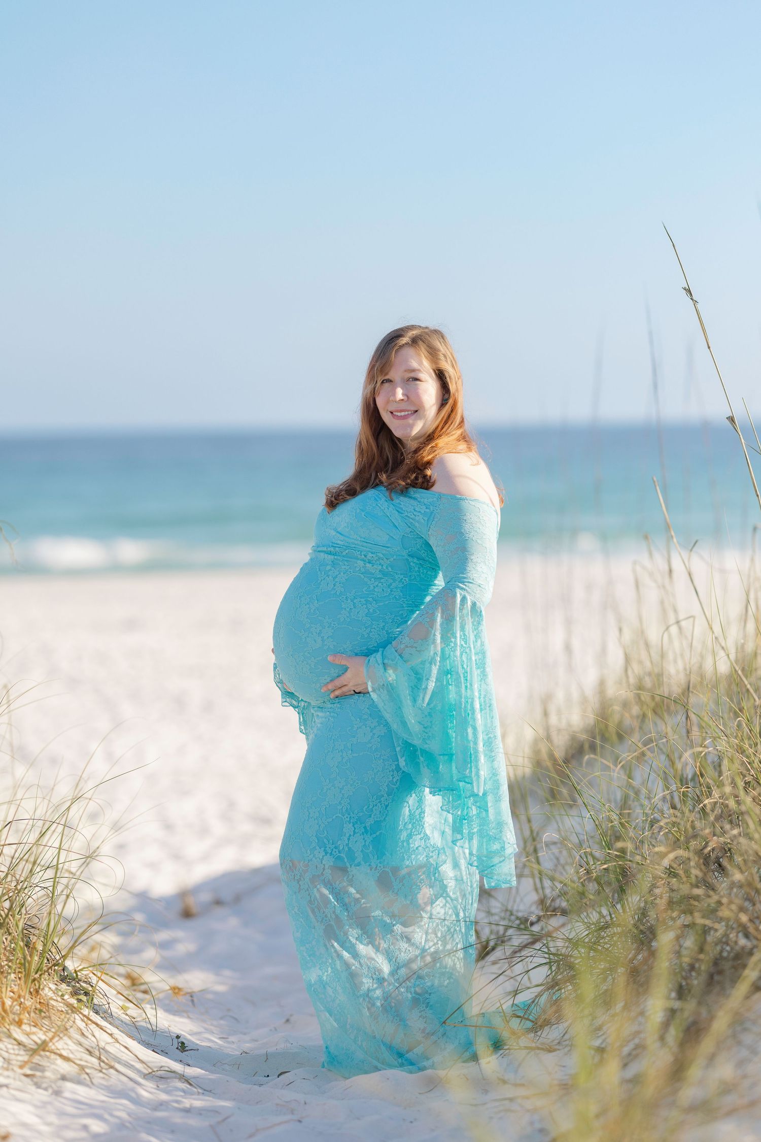 Pregnant woman in teal lace gown standing on white sand beach dunes, cradling her baby bump with ocean waves behind her.