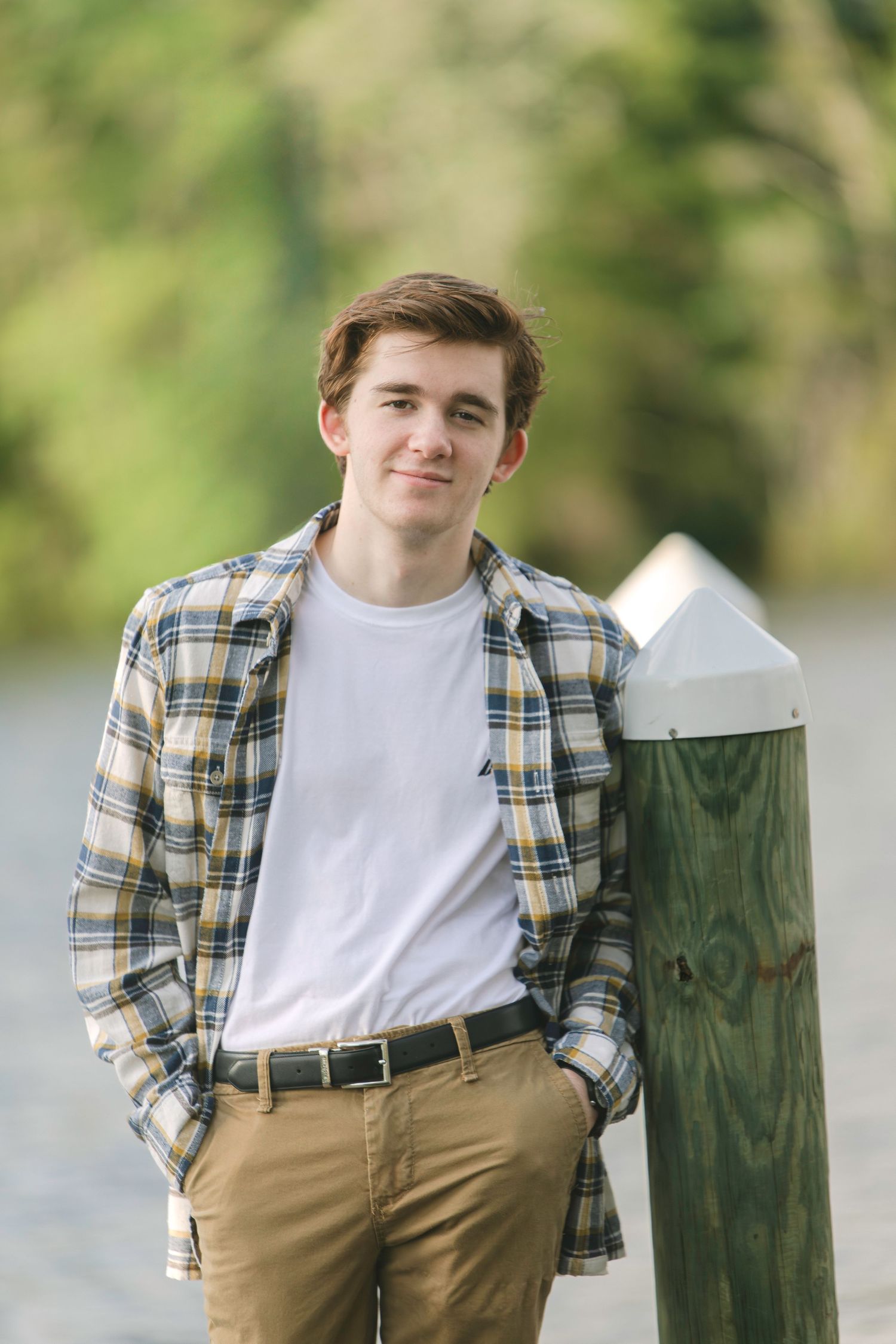Young man in plaid shirt and khakis leaning against a wooden dock post outdoors.