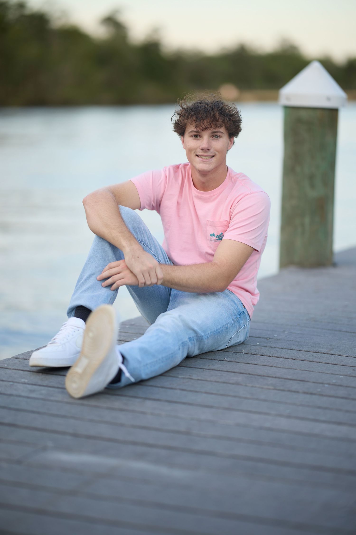 Young man in pink shirt and jeans sitting on a wooden dock by the water, smiling at the camera.