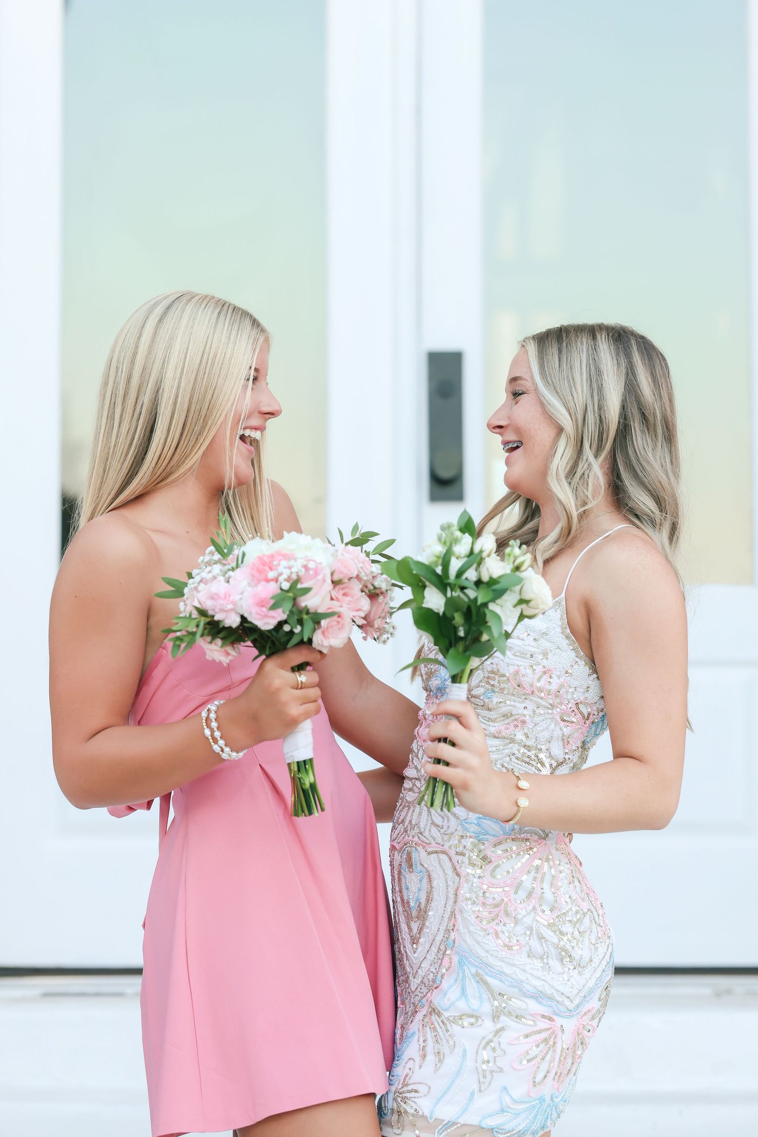 Two smiling blonde women in pink and floral dresses holding bouquets of pink flowers in front of white doors.