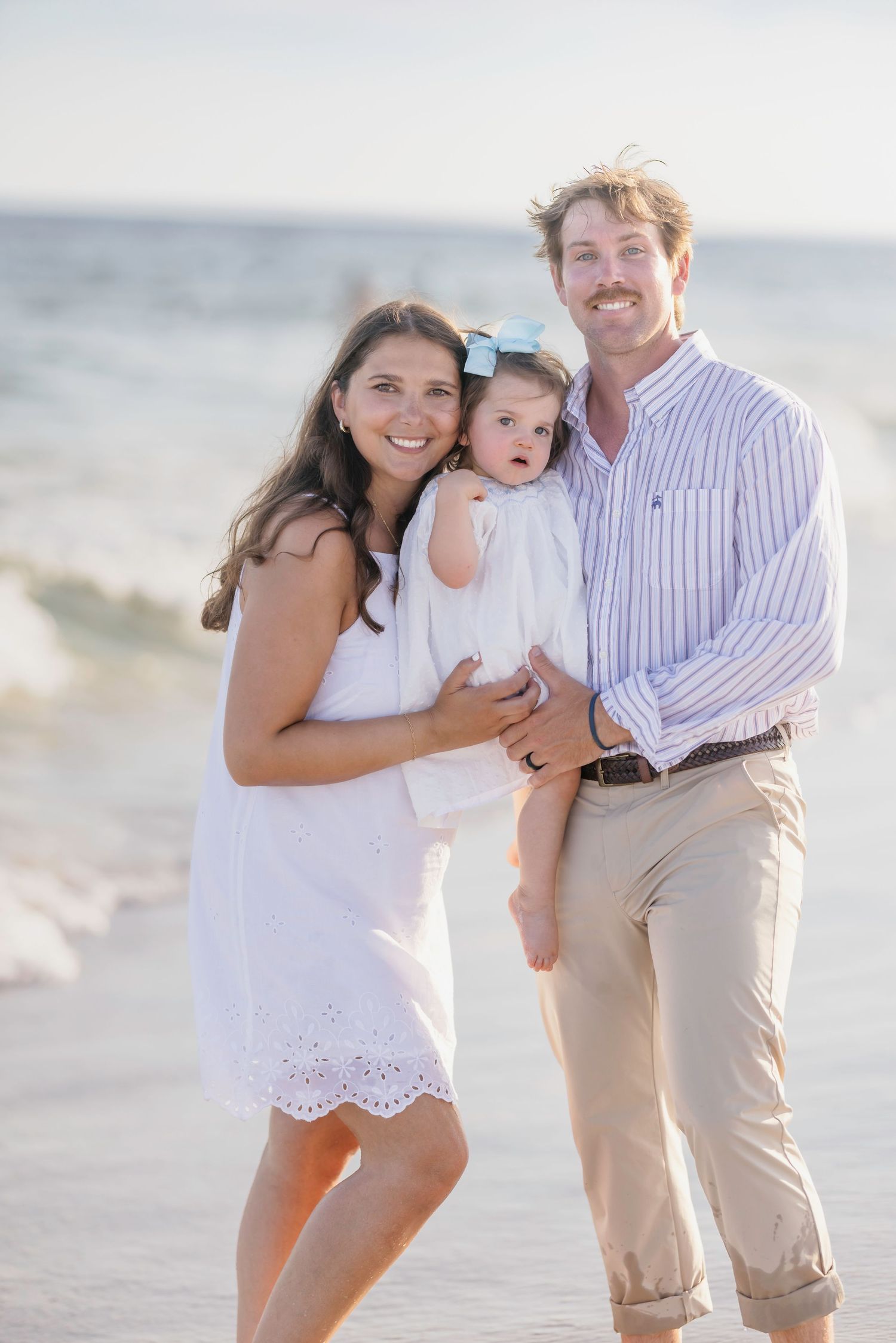 Happy family of three posing on beach with waves, parents holding baby girl.
