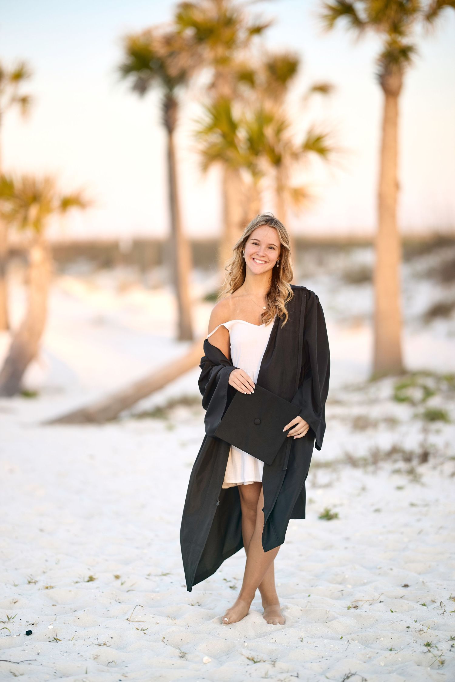 Blonde woman in white dress holding black graduation cap and gown on sandy beach with palm trees at sunset.