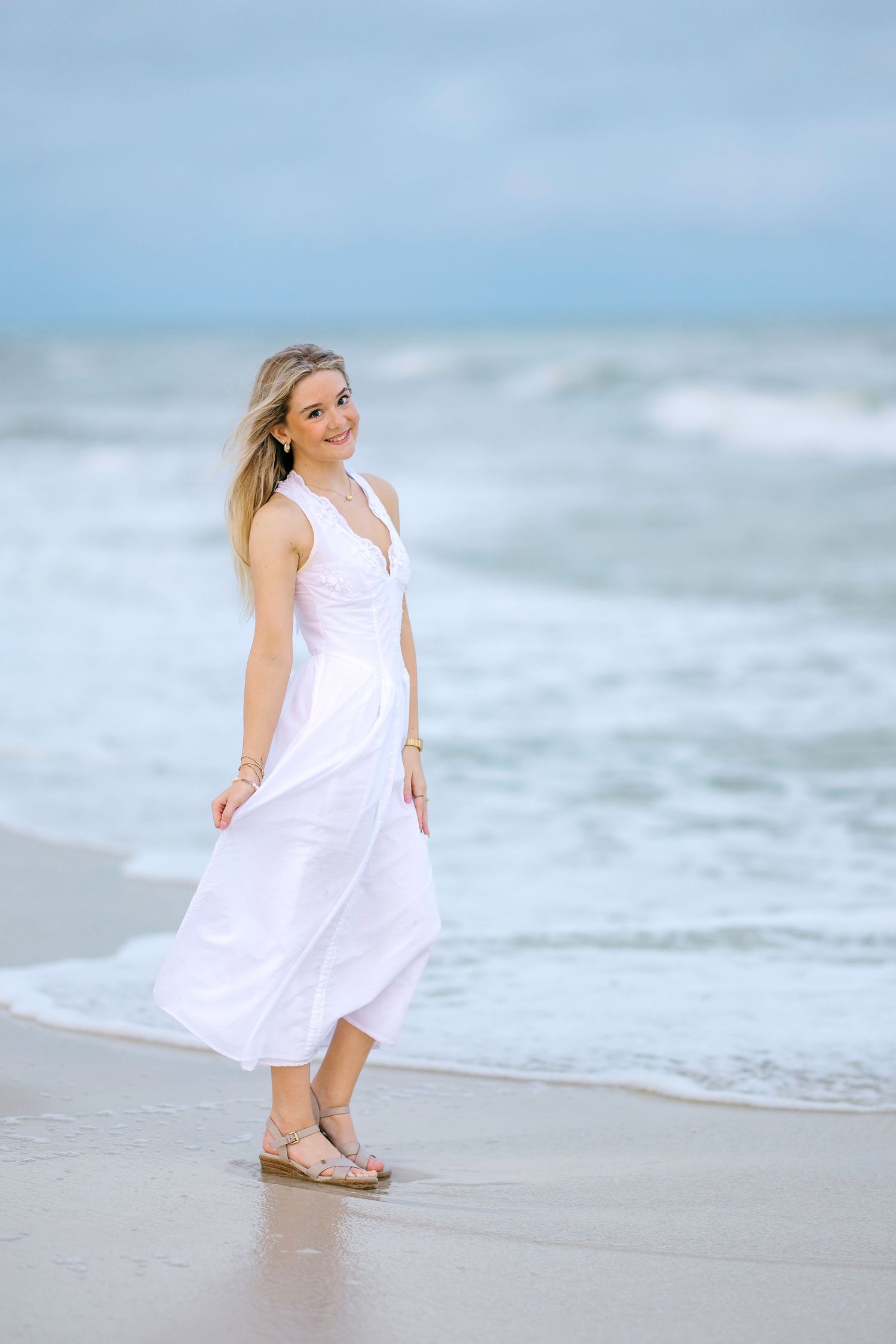 A smiling blonde woman in a flowing white maxi dress walks along a sandy beach with ocean waves behind her.