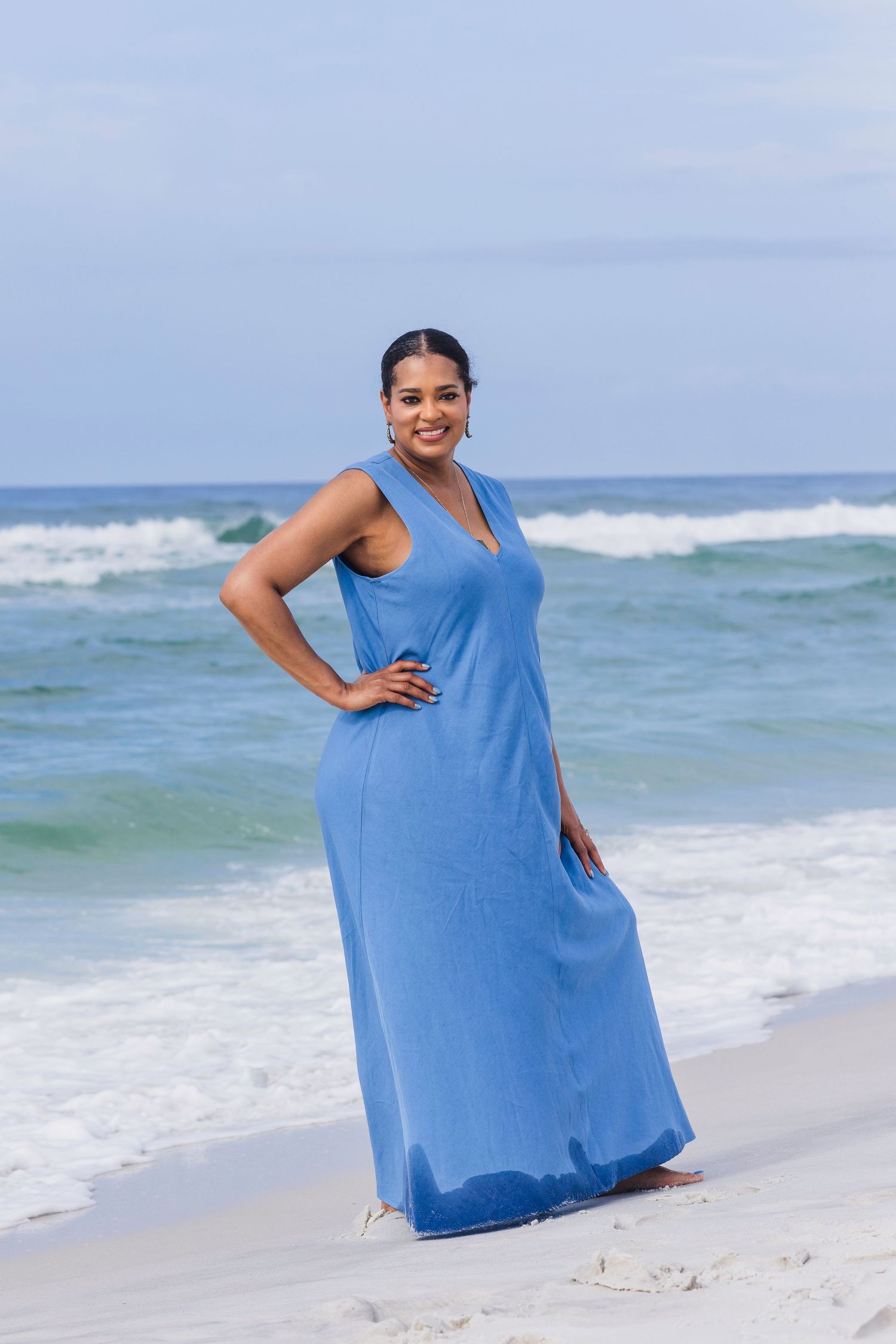 Woman in elegant blue maxi dress posing confidently on sandy beach with ocean waves in the background.