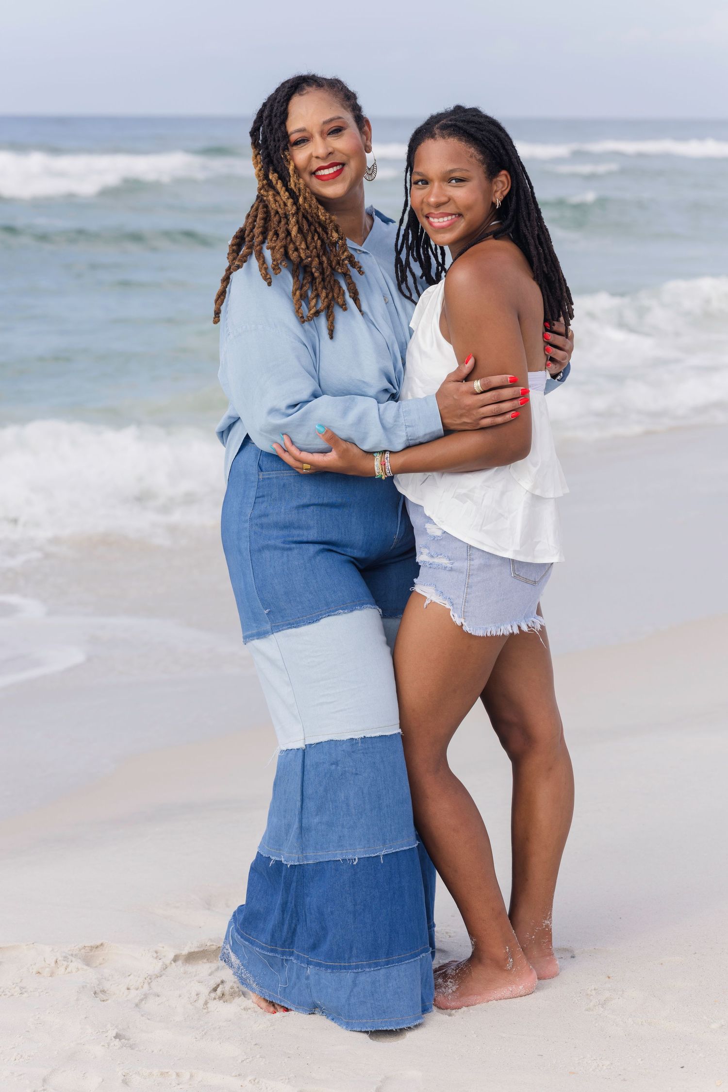 Two women with braided locs share a warm embrace on a sunny beach with ocean waves in the background.