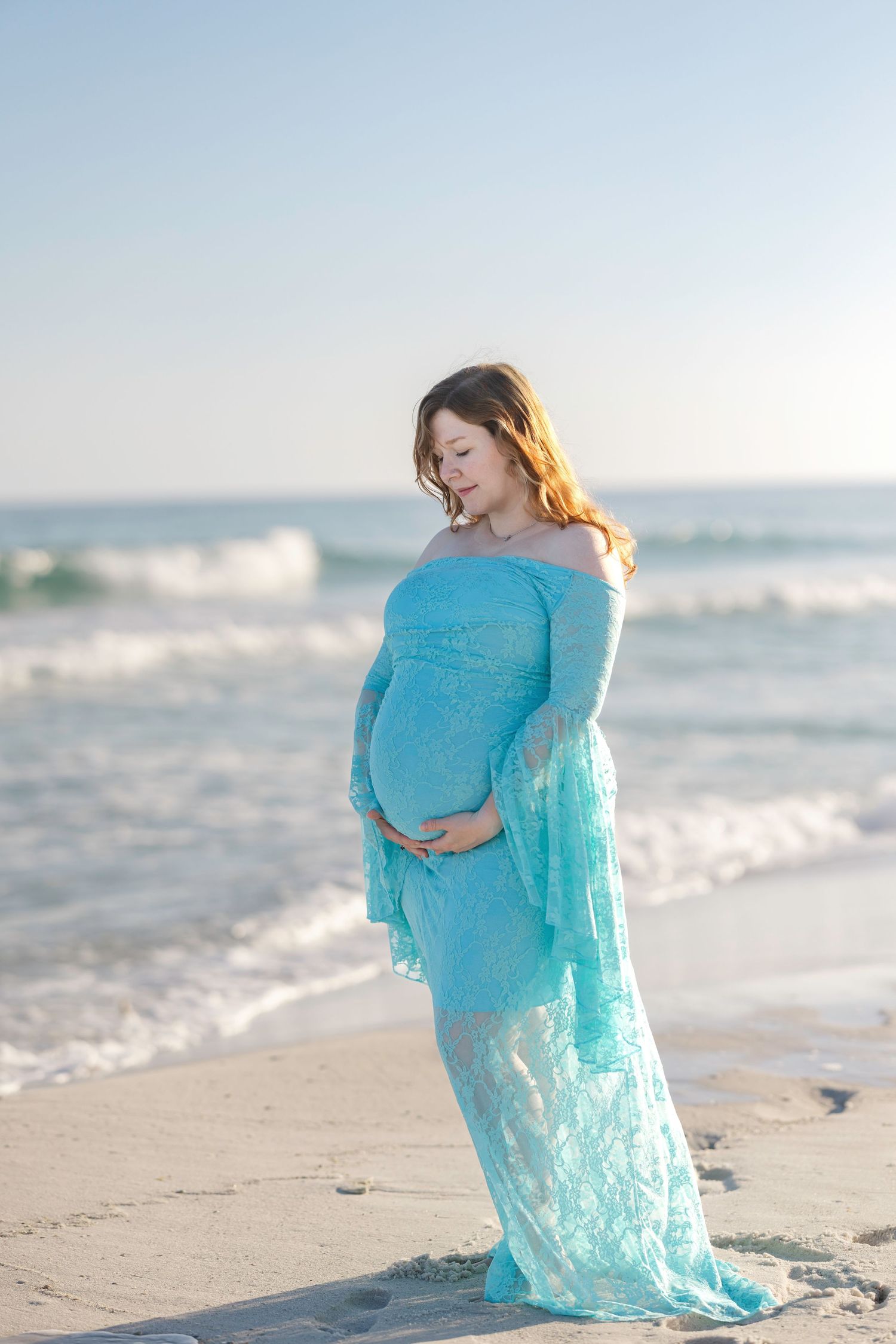 Pregnant woman in a flowing teal lace gown standing on a sandy beach, cradling her baby bump with ocean waves behind her.