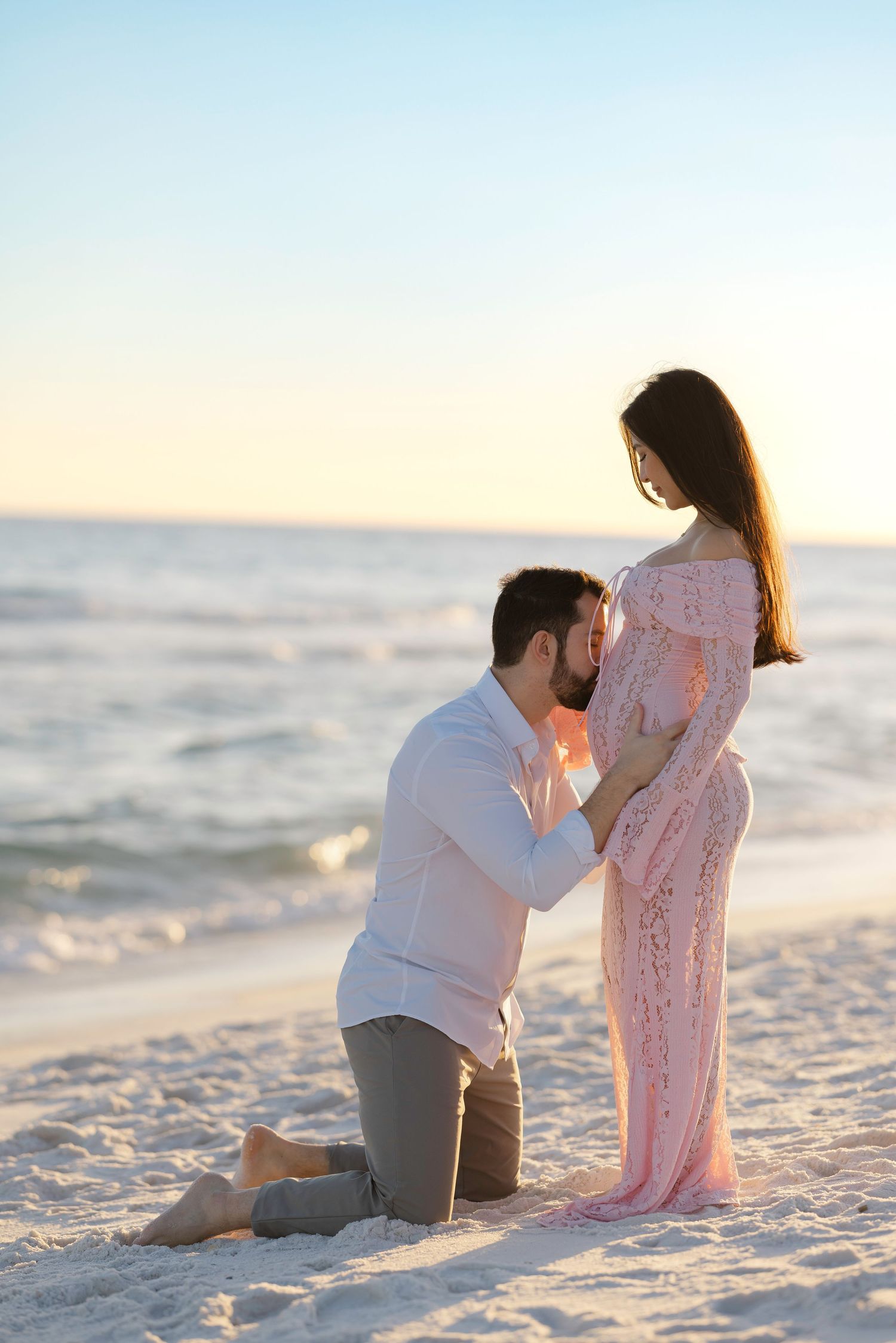 Man kneeling on beach kissing pregnant woman's belly at sunset, romantic maternity photo.