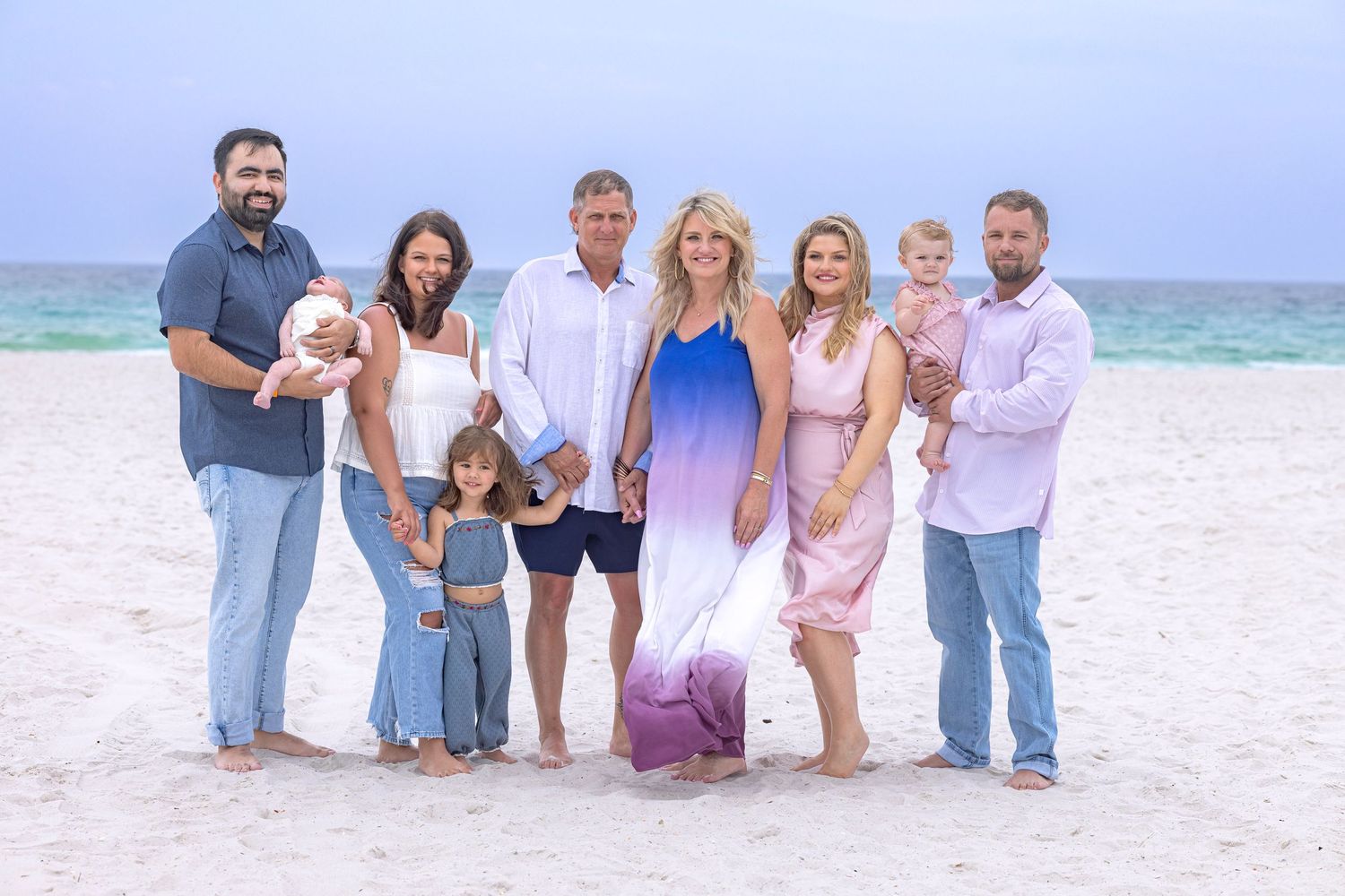 A large family poses together on a sandy beach with turquoise water, including adults and young children in coordinated outfits.