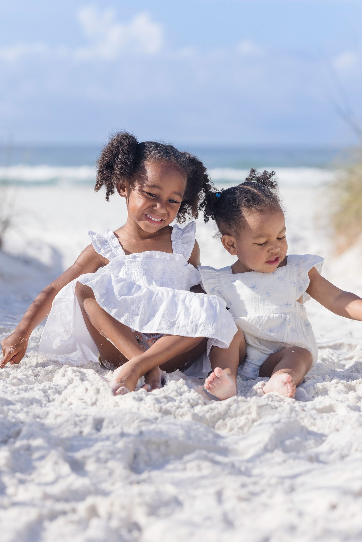 Two young African American girls in white dresses playing in white sand at the beach on a sunny day.
