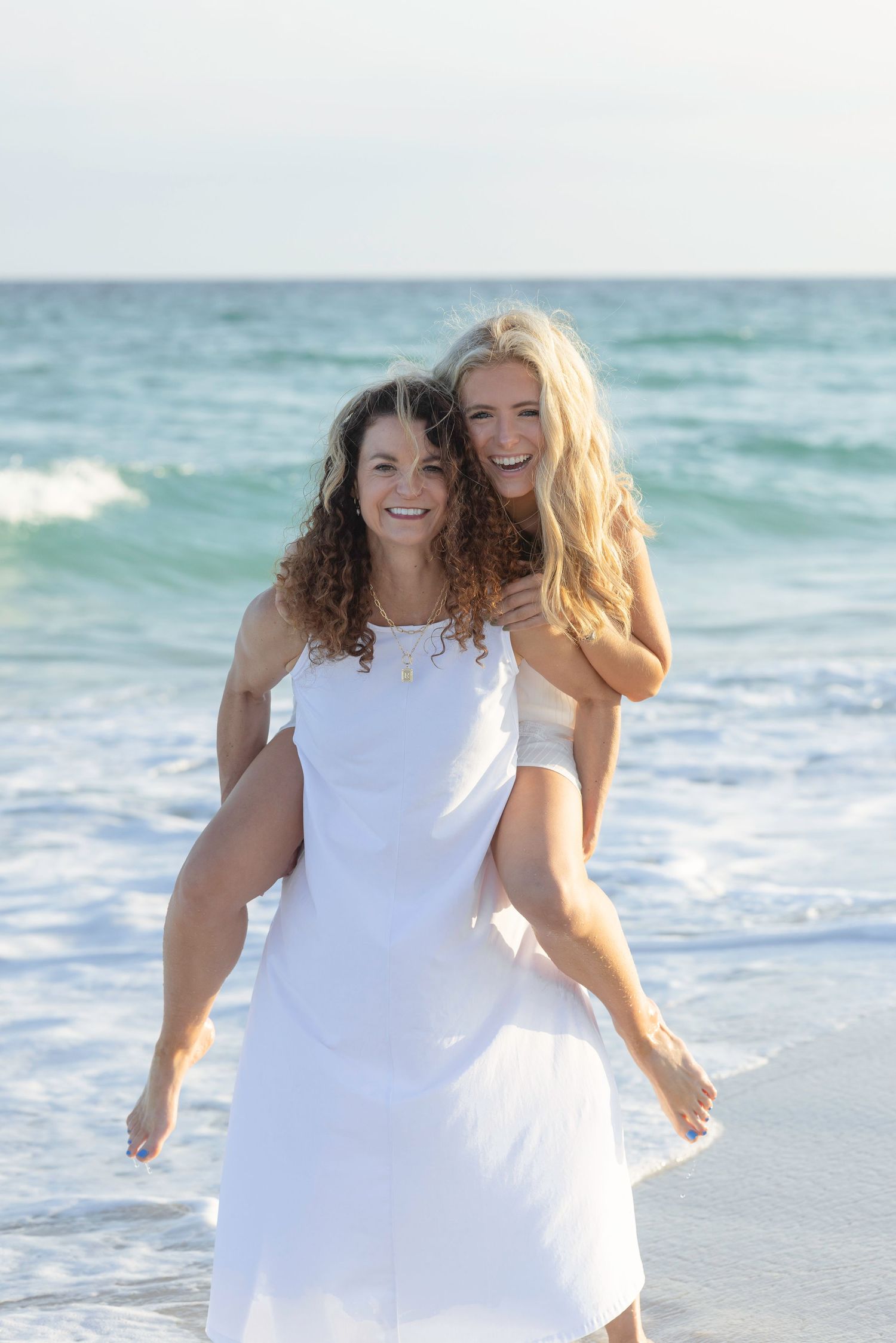 Two happy women in white dresses laughing on a beach, one giving the other a piggyback ride.
