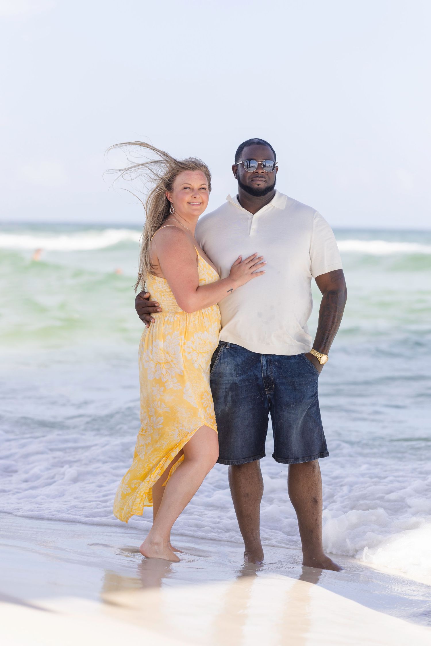 Smiling interracial couple embracing on a sunny beach with ocean waves, woman in yellow dress, man in white shirt.