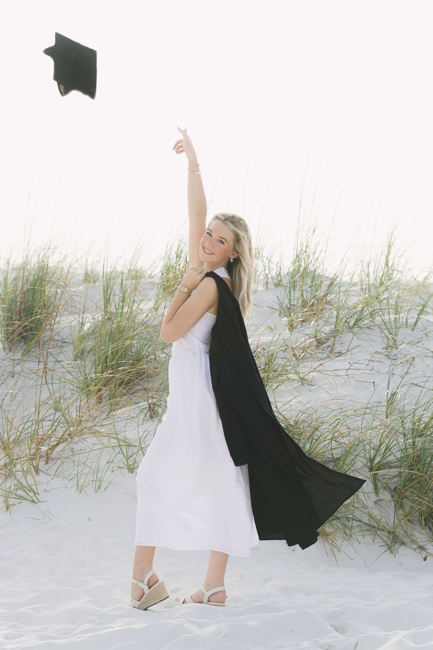 Graduate in white dress and black graduation gown throws cap on beach with sea grass in background.