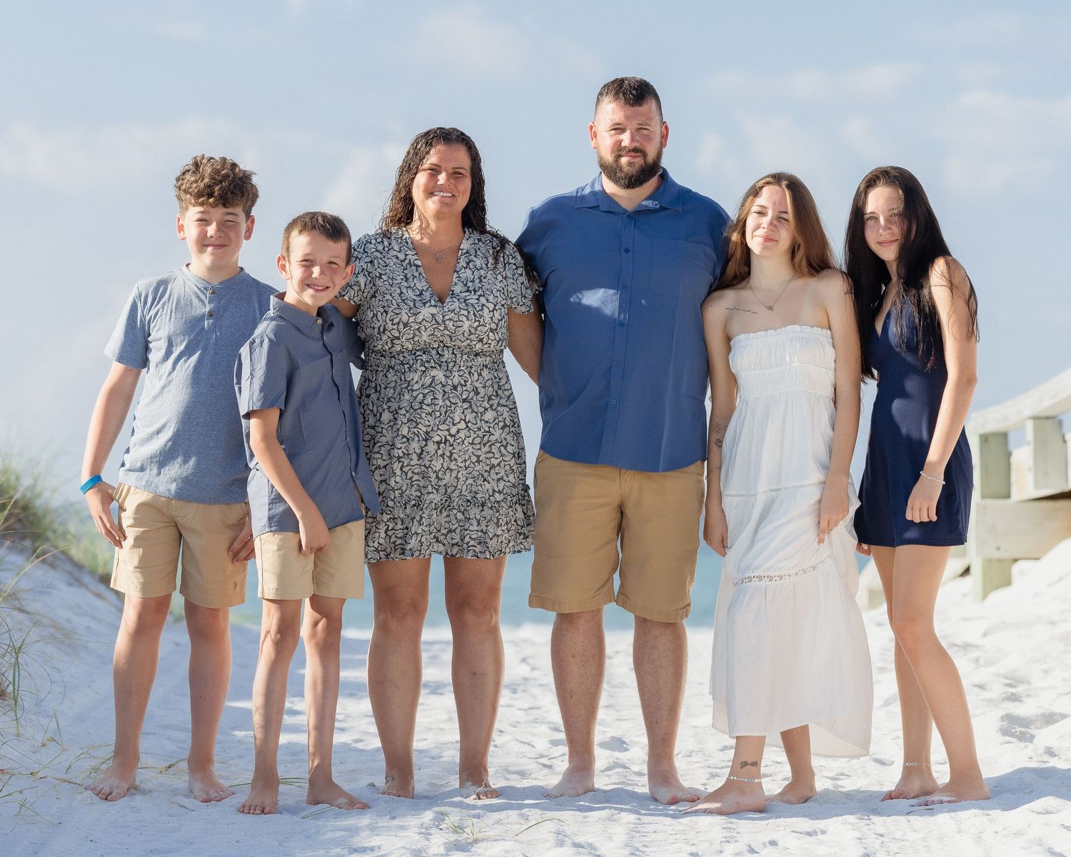 Family of six posing together on a sandy beach with blue skies, wearing casual summer outfits.