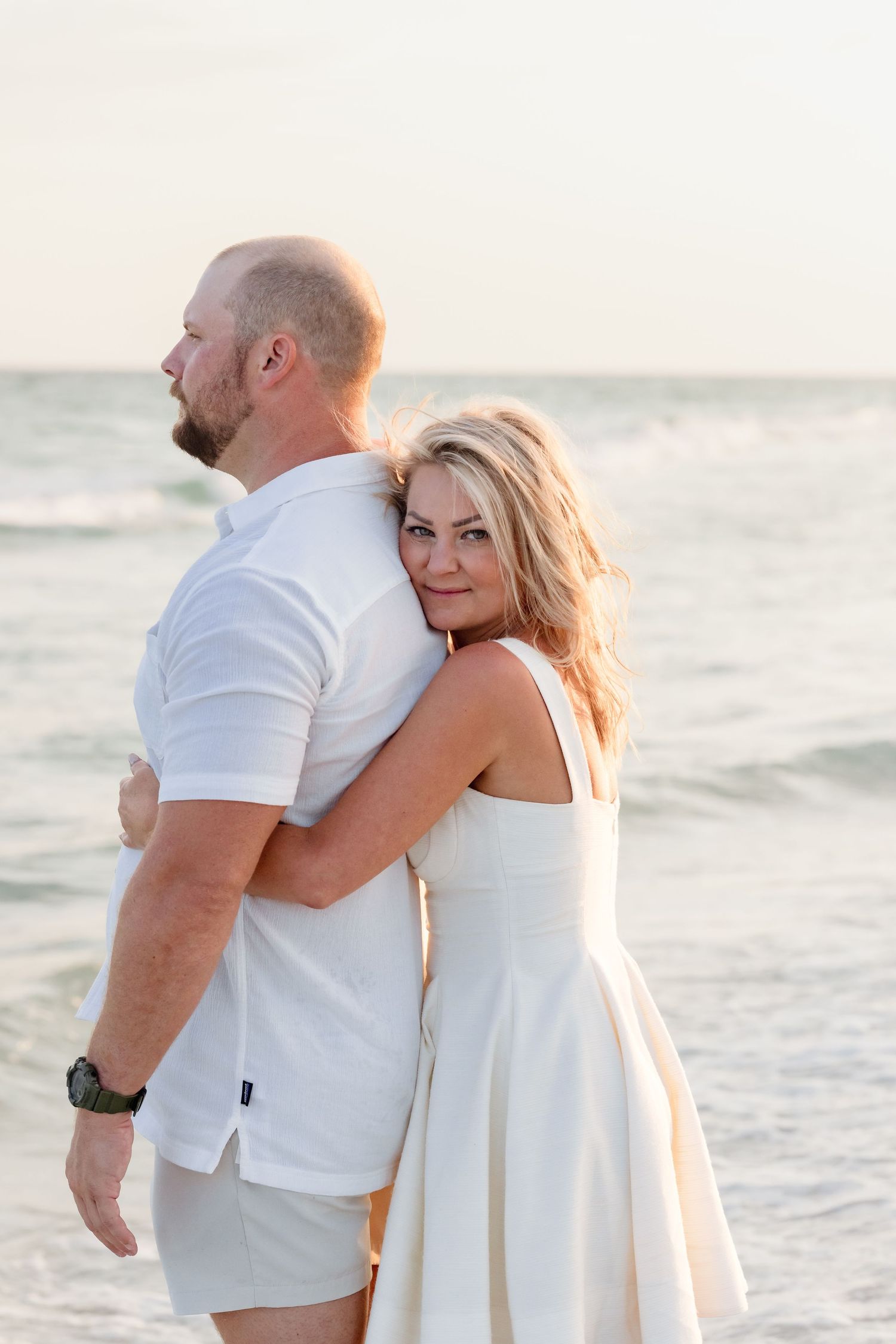 Romantic couple in white outfits embracing on a beach with ocean waves behind them.