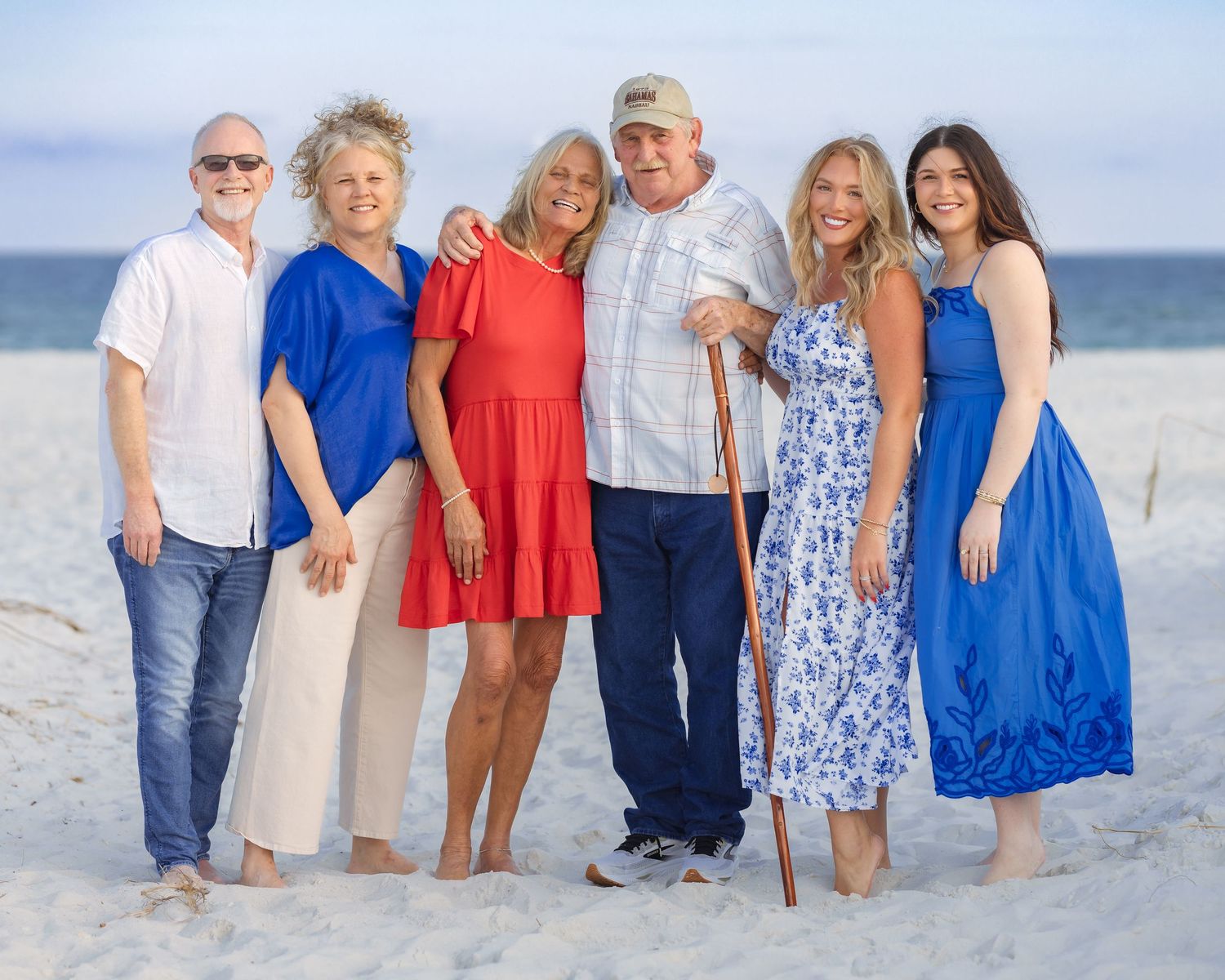 Family of six smiling together on a sandy beach, dressed in coordinating blue, red, and white outfits.