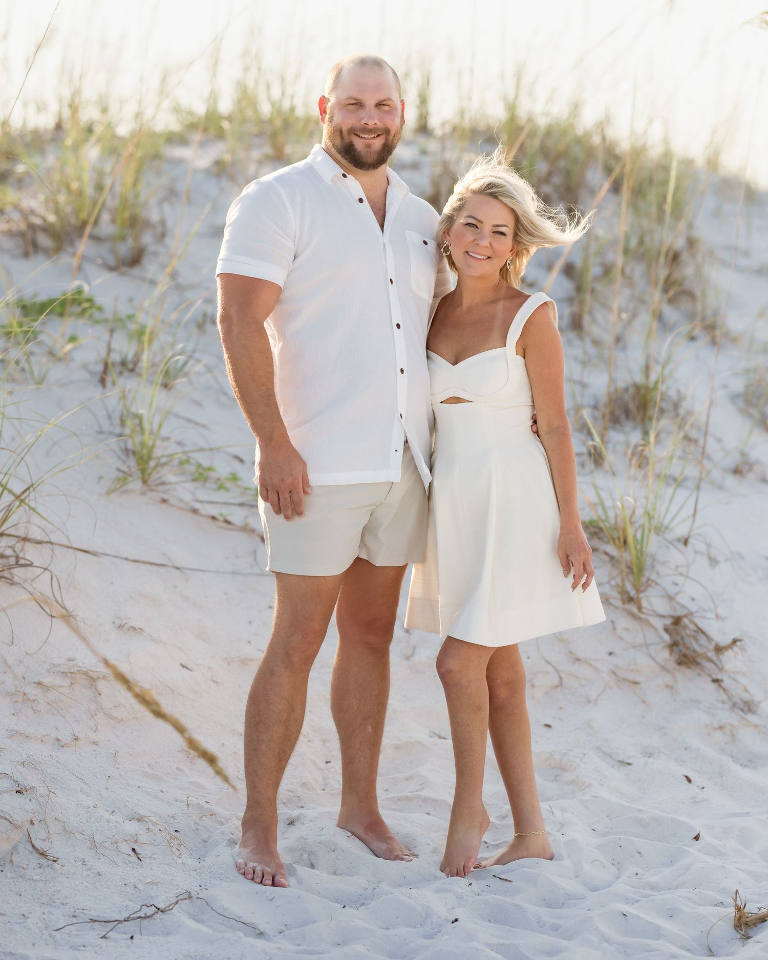 Couple in matching white outfits posing barefoot on sandy beach dunes with sea grass in background.