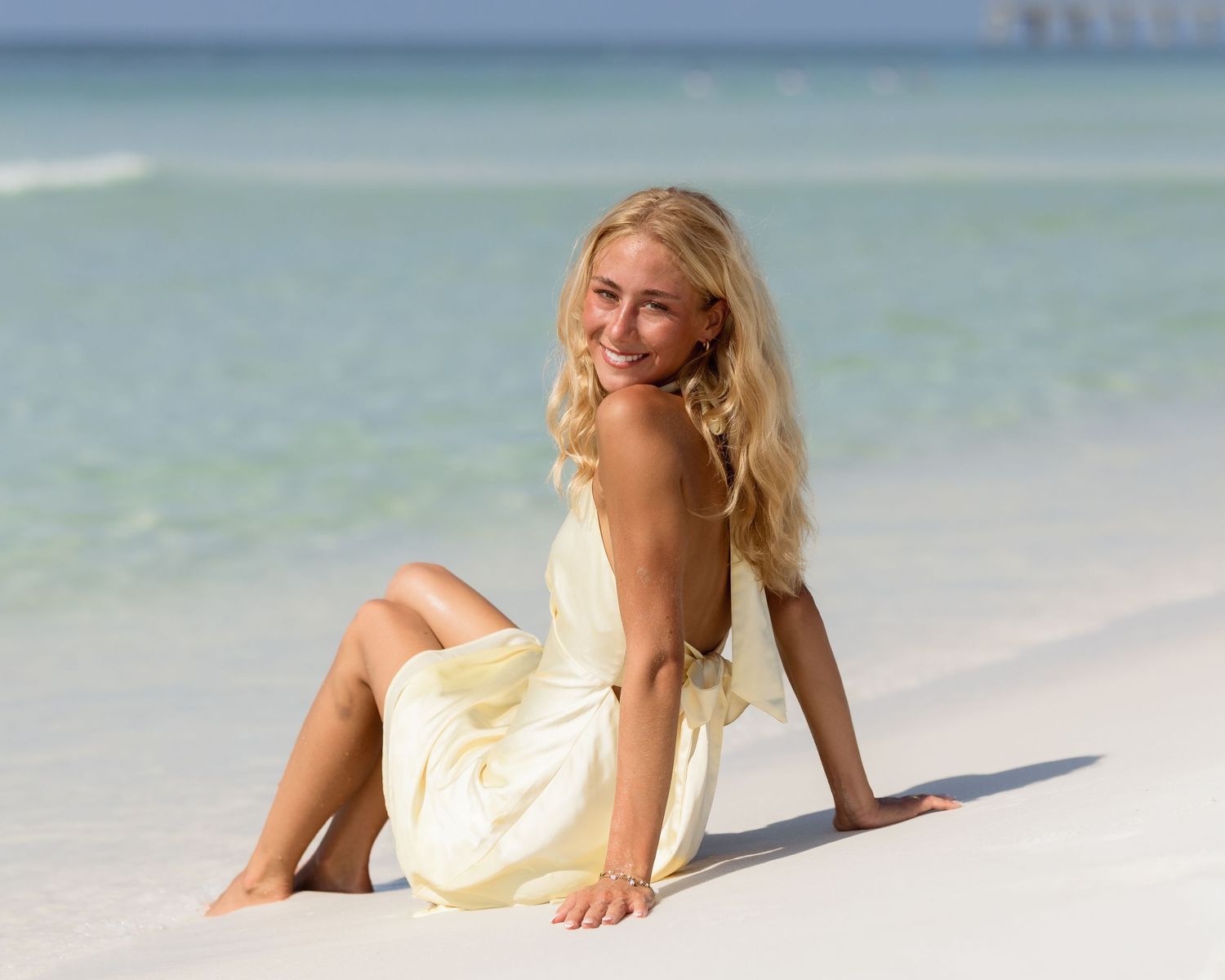 Blonde woman in yellow dress sitting on white sandy beach.