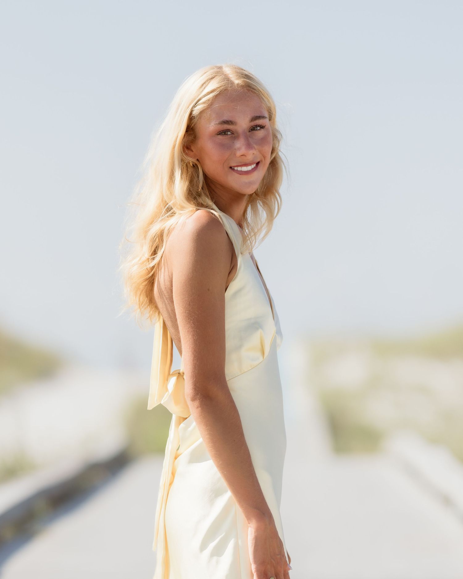 Young blonde woman in a light yellow dress smiling outdoors on a sunny beach path.