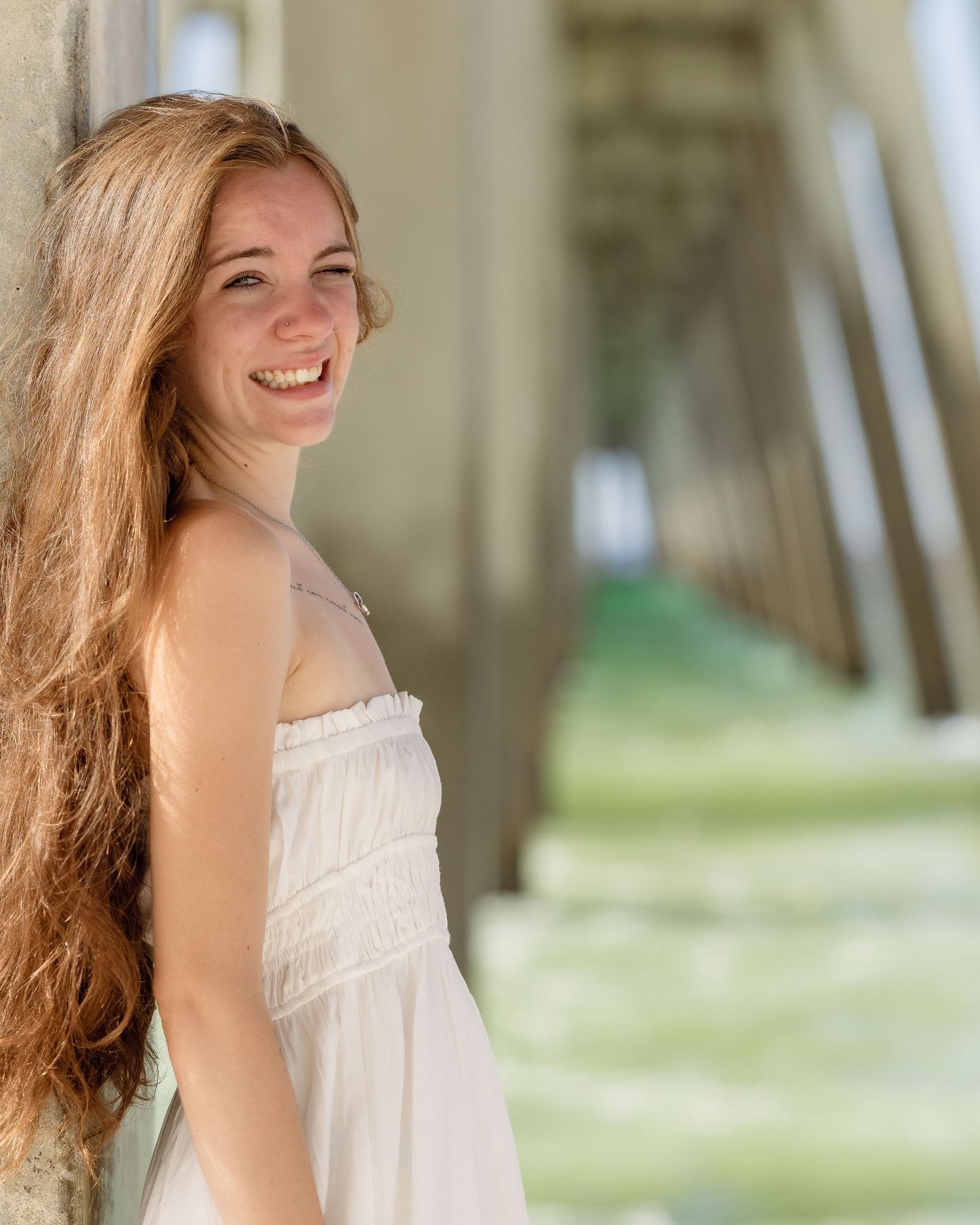 Smiling young woman in white sundress leaning against pier column with long red hair.