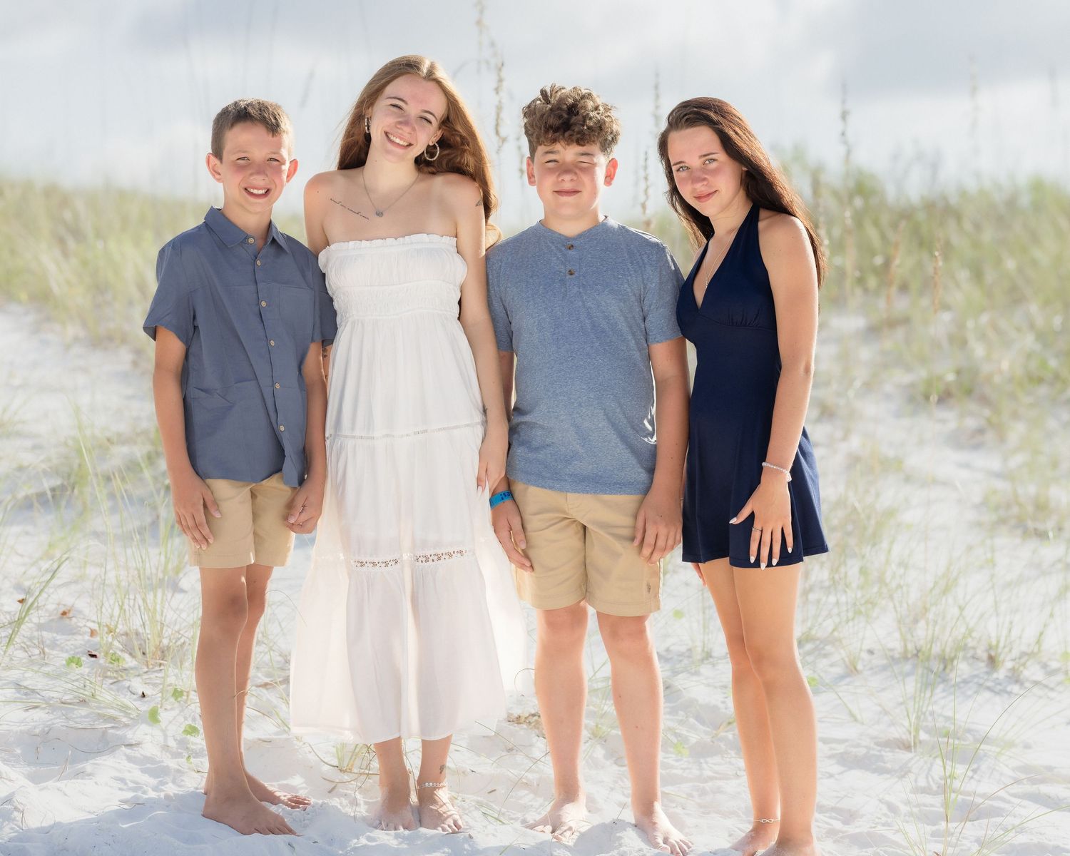 Four teenagers smiling together on a sandy beach with dunes and sea oats in the background on a sunny day.