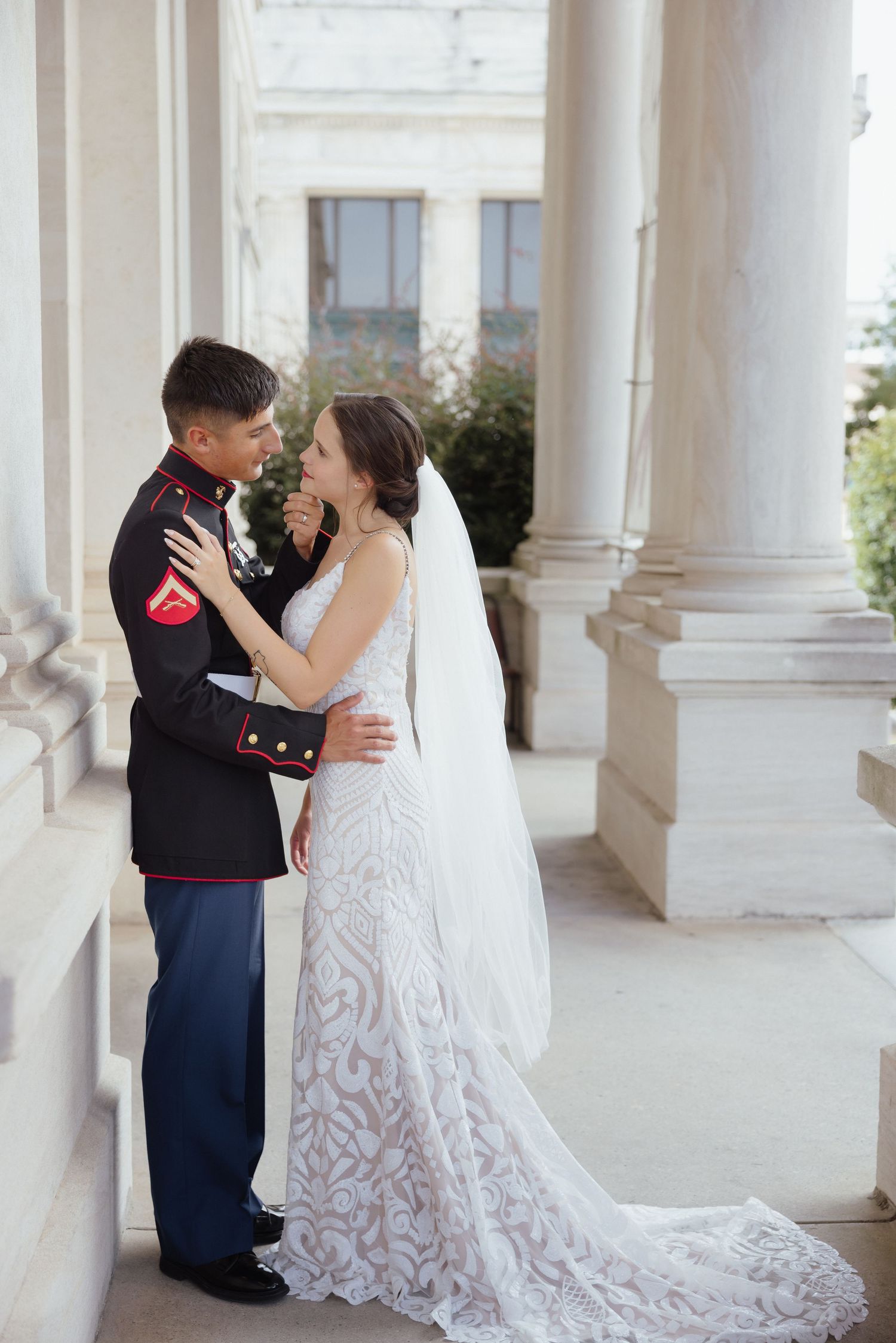 Marine groom in dress uniform embraces bride in lace wedding gown with veil between white columns.
