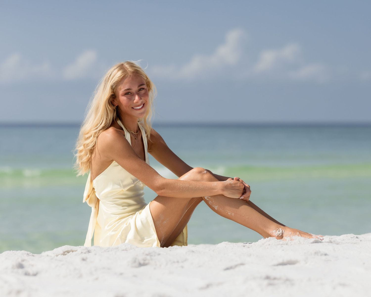 Smiling blonde woman in yellow dress sitting on white sand beach with turquoise ocean in background.