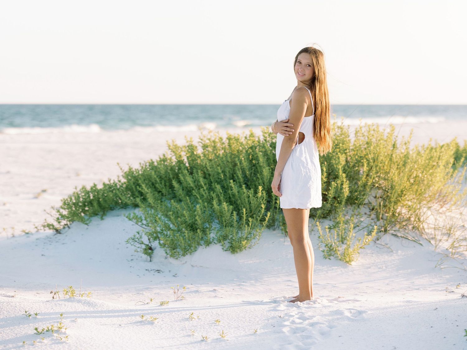 Person in white dress stands on beach dunes against ocean backdrop.