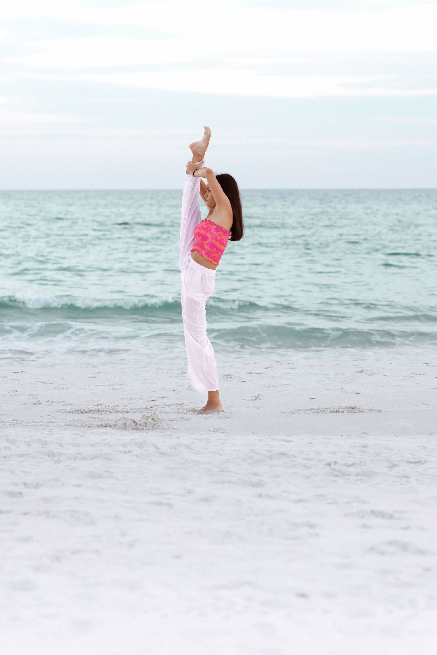 Person in pink top and white pants dances joyfully on beach with waves in background.