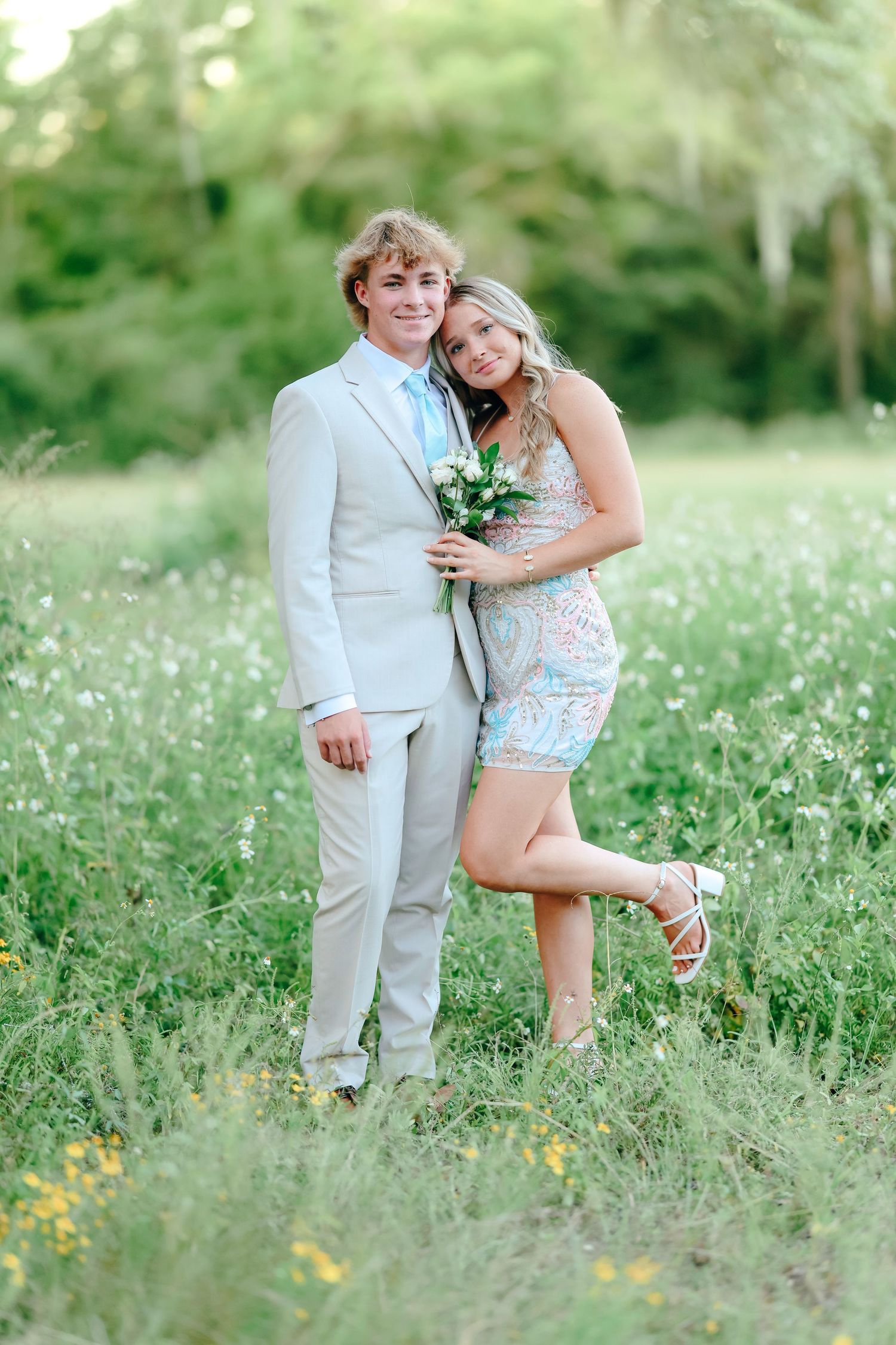 A young couple poses in a flower field, the man in a light suit and the woman in a floral dress holding a bouquet.
