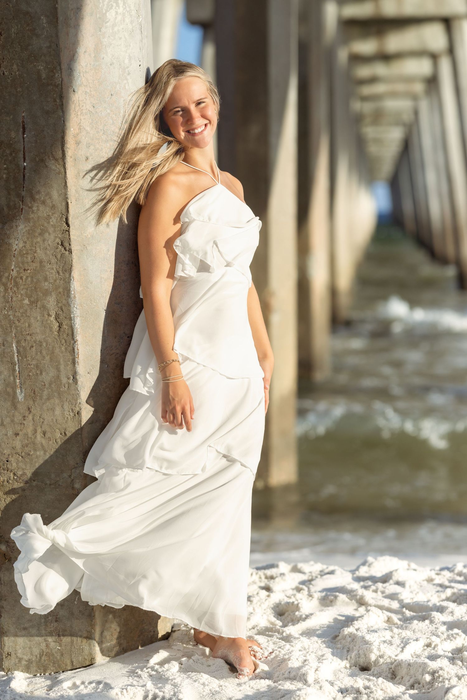 Smiling blonde woman in flowing white dress leaning against pier pillar on sandy beach.