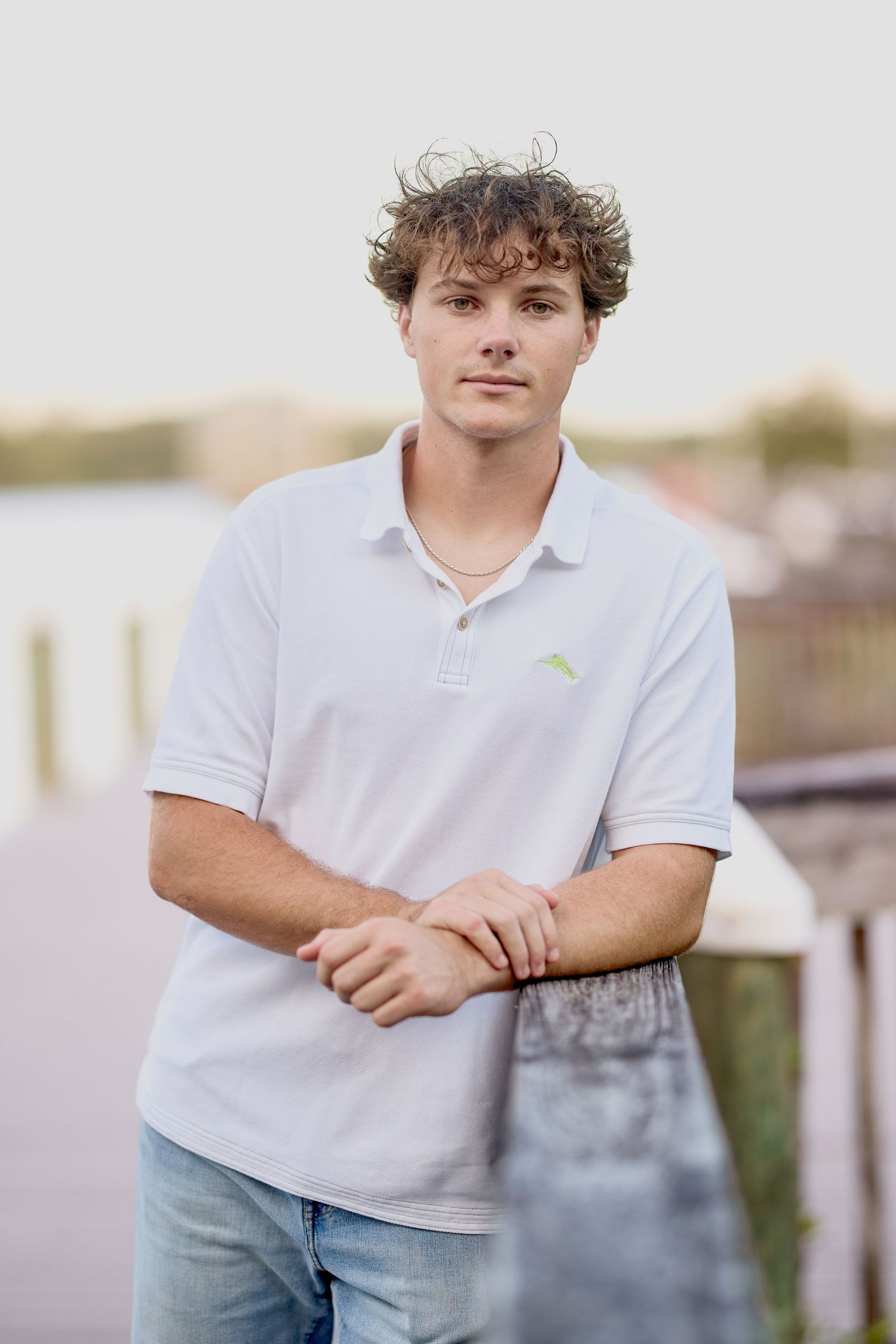 Young man in white polo shirt poses near waterfront dock with blurred marina background.