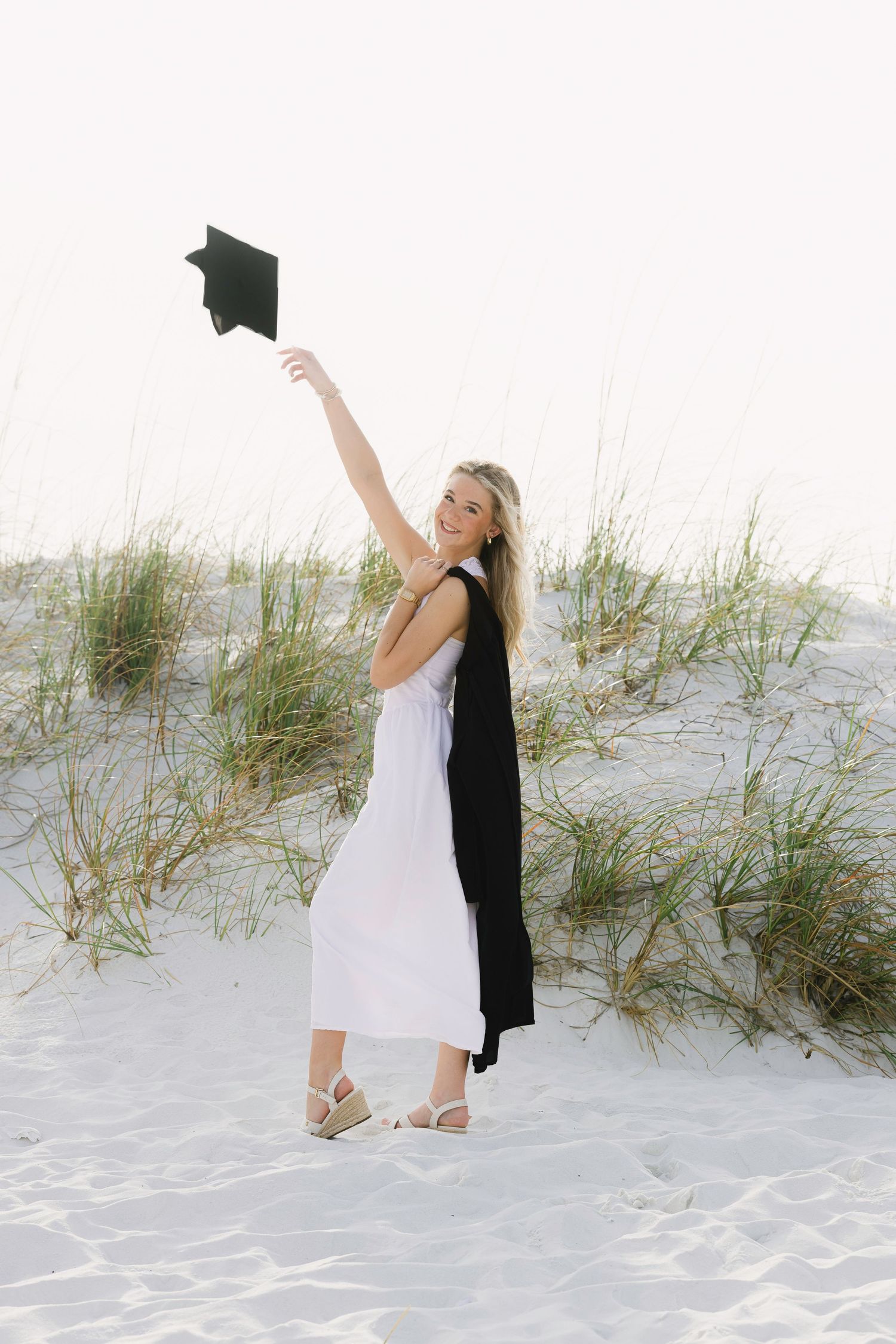 Graduate in white dress and black robe celebrates on beach sand dunes while holding up graduation cap.