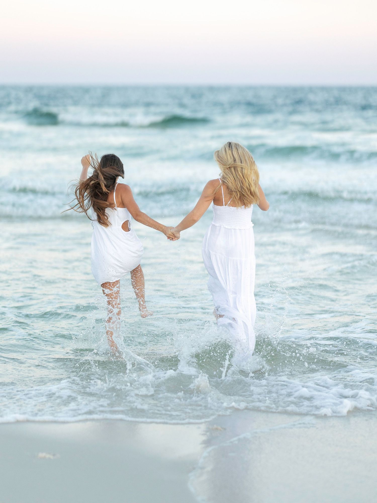 Two people in white dresses splash and play together in the ocean waves at sunset.