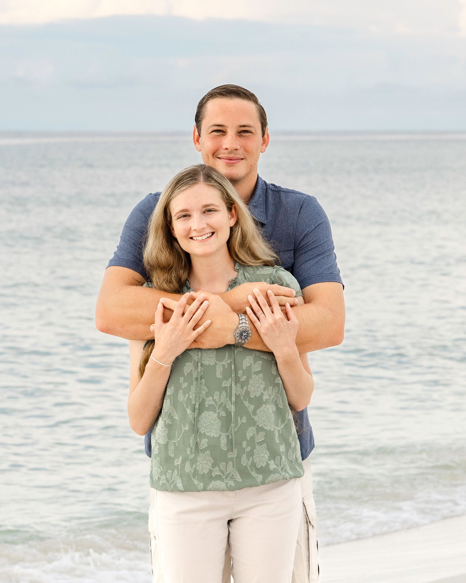 A couple embraces on a sunny beach with gentle waves and soft sand in the background.