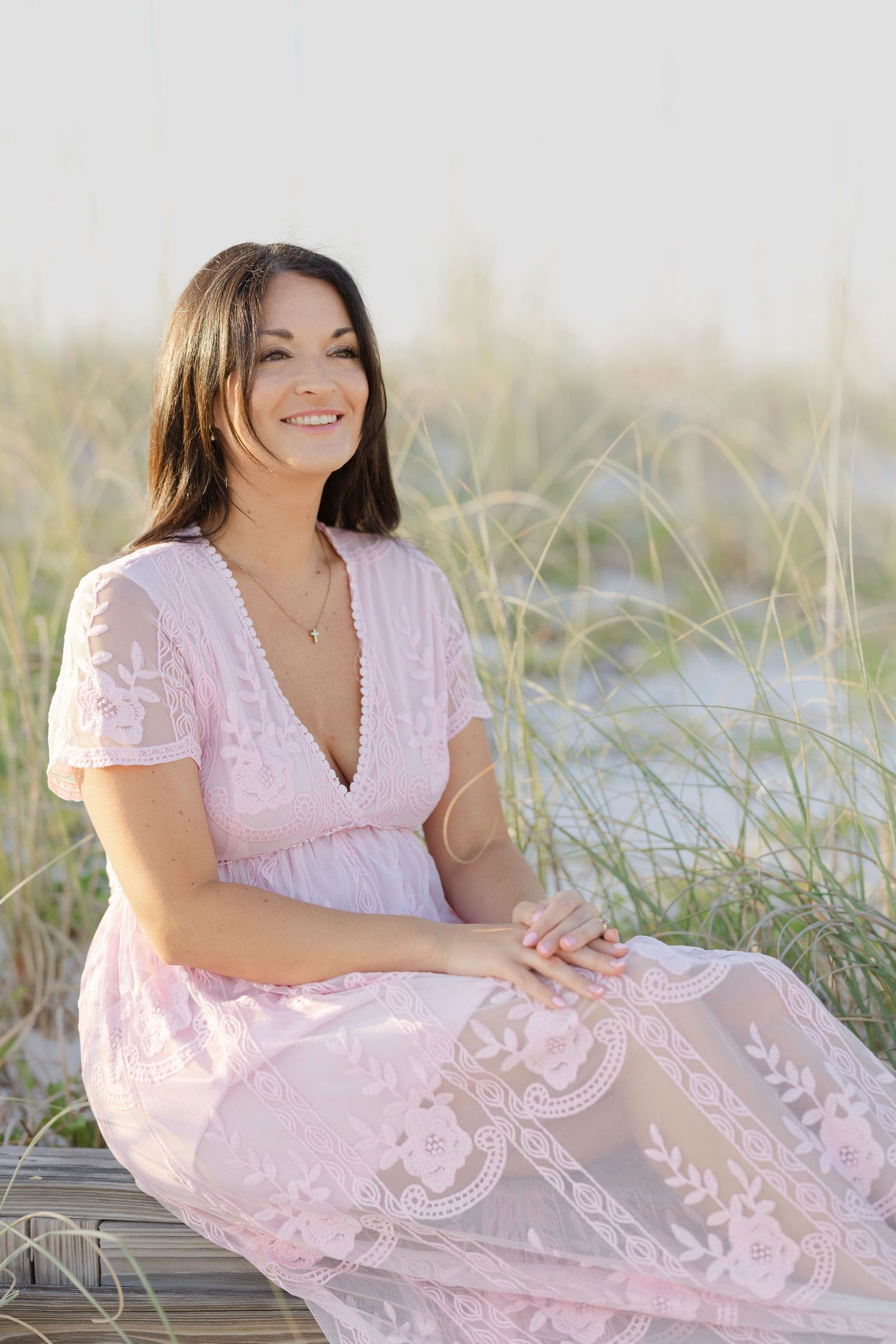 A person in a soft pink lace dress sits gracefully among beach grass during a coastal outdoor photoshoot.