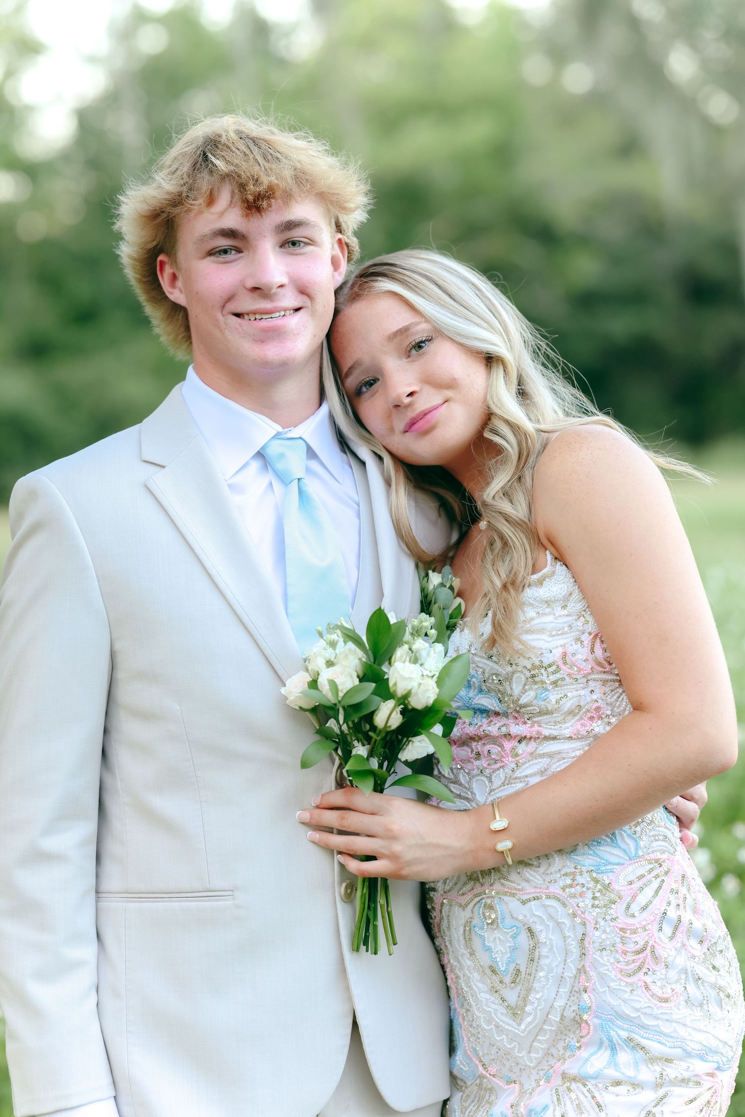 A couple dressed in formal attire posing together outdoors while holding a white floral bouquet.