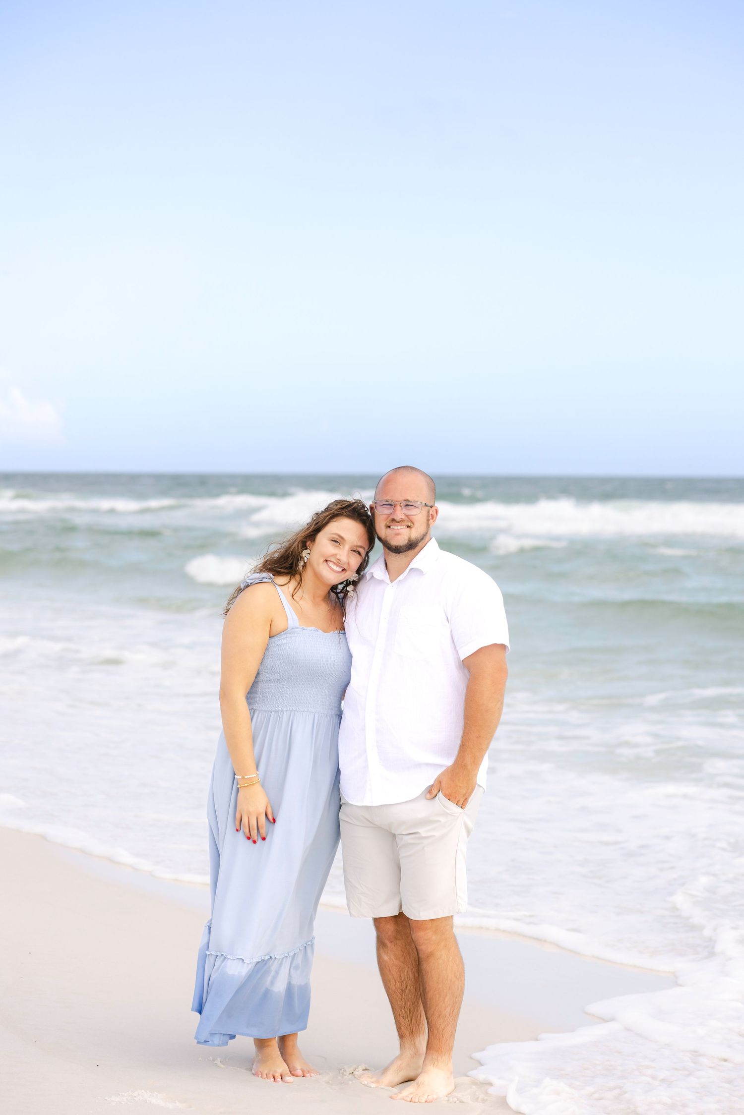 Couple in light blue and white outfits sharing intimate moment on beach with waves crashing behind.