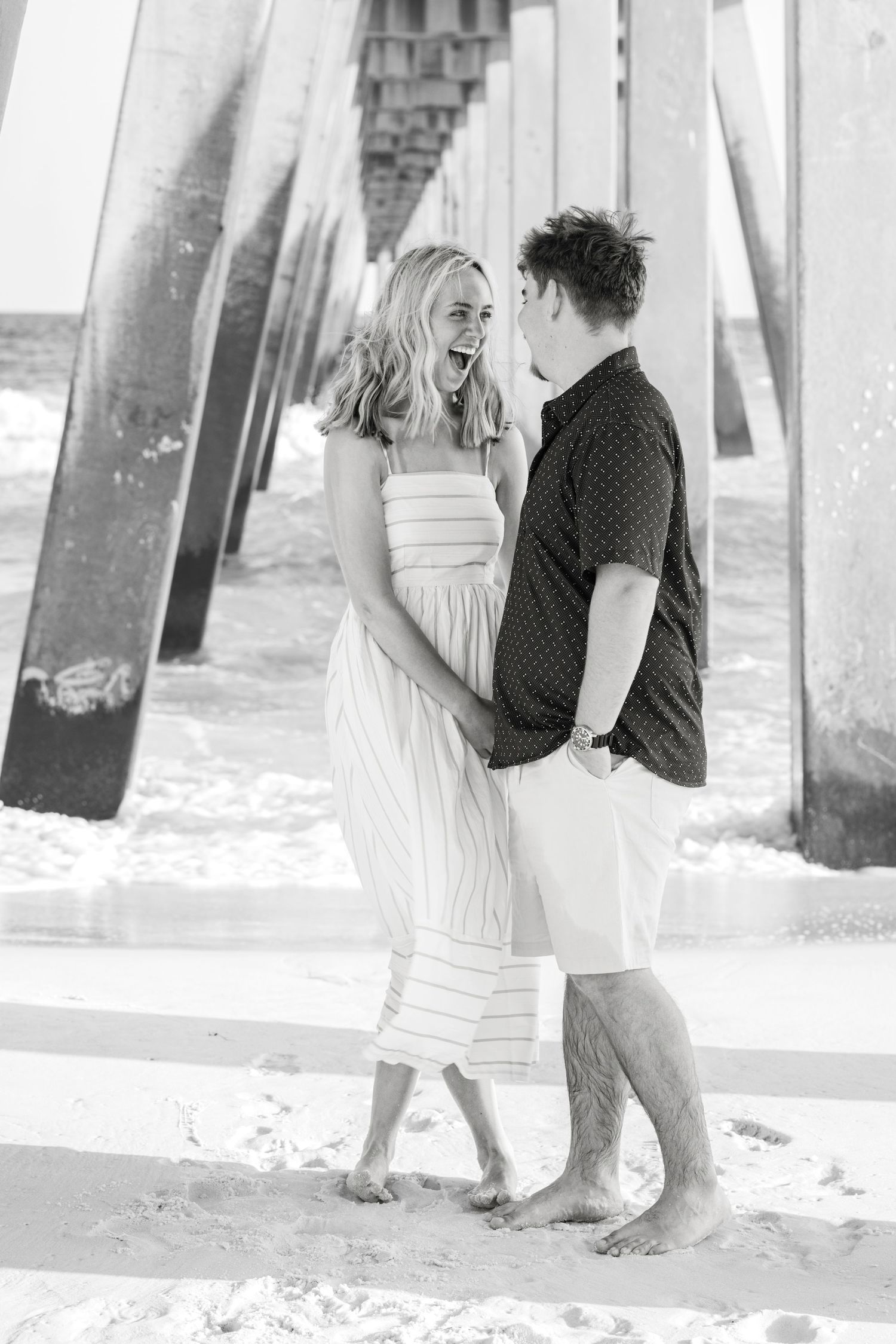Black and white photo of a laughing couple embracing barefoot on sandy beach under a wooden pier.