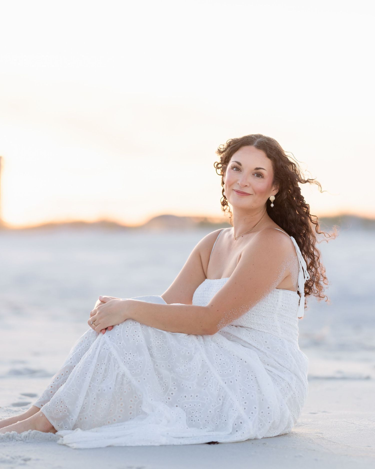 Woman in white eyelet dress sitting on sandy beach smiling at golden sunset light.