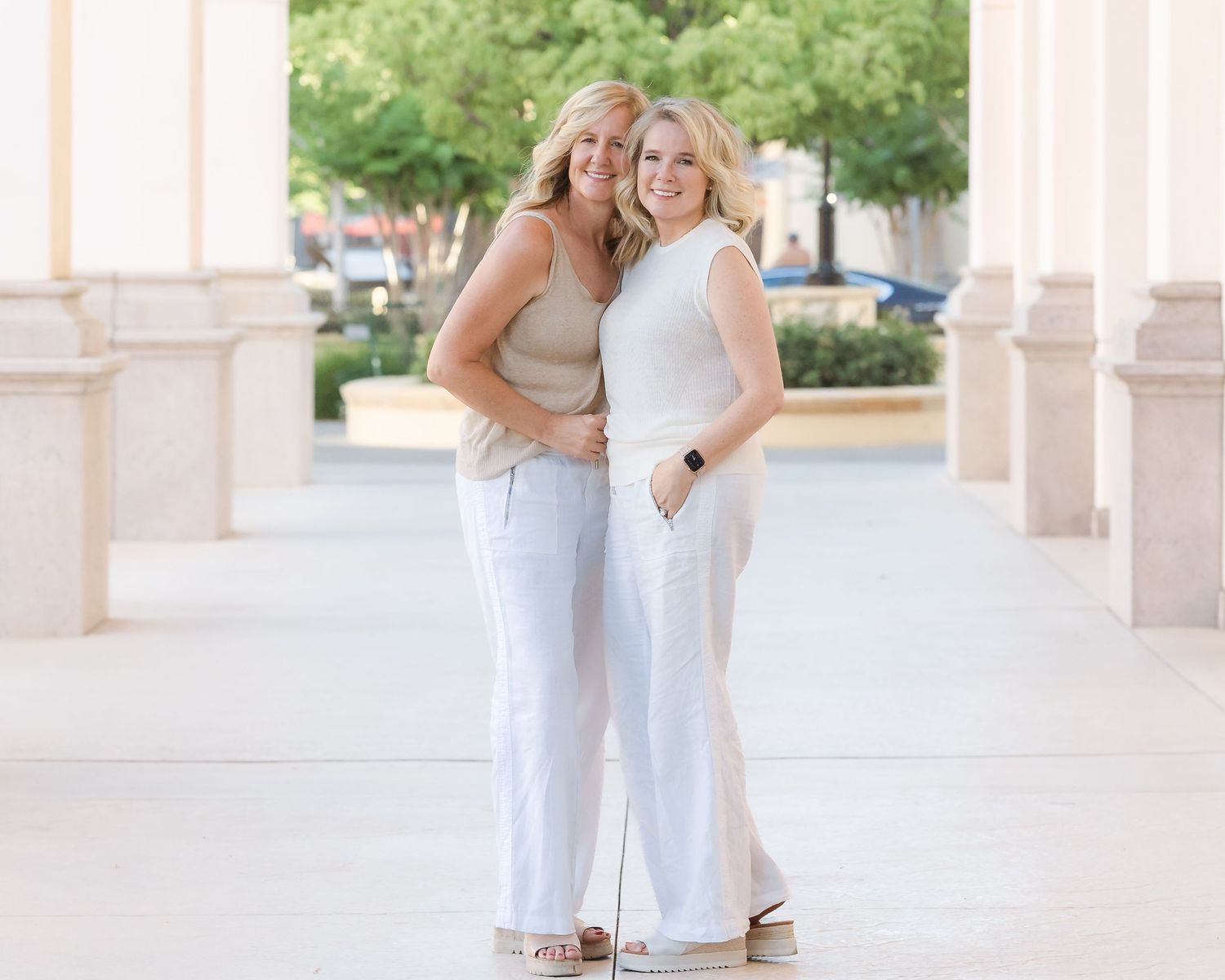 Two smiling blonde women posing together outdoors near columns, wearing white pants and neutral tops.