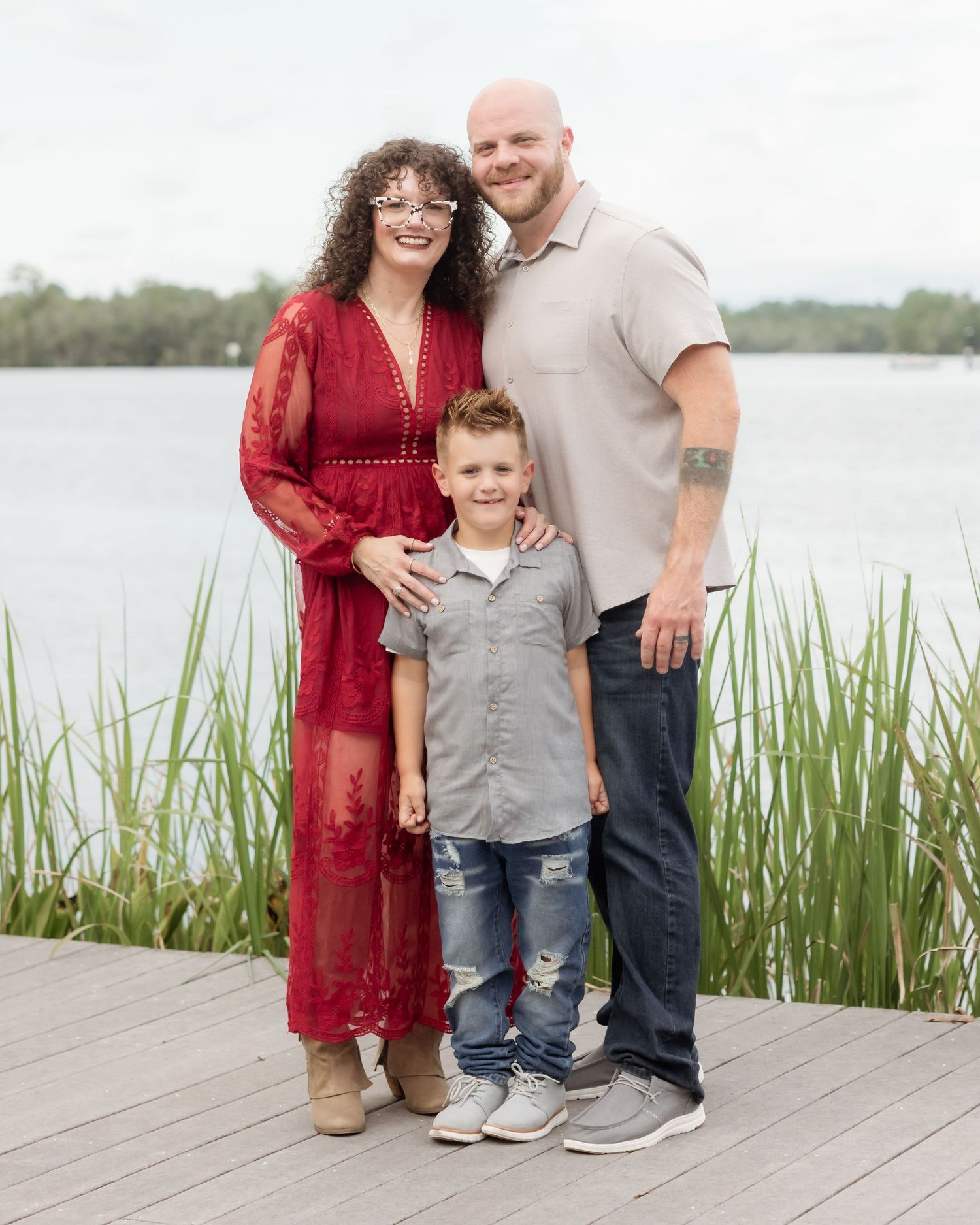 Family of three posing on a dock by a lake, woman in red dress, bald man with tattoo, and young boy in gray shirt.