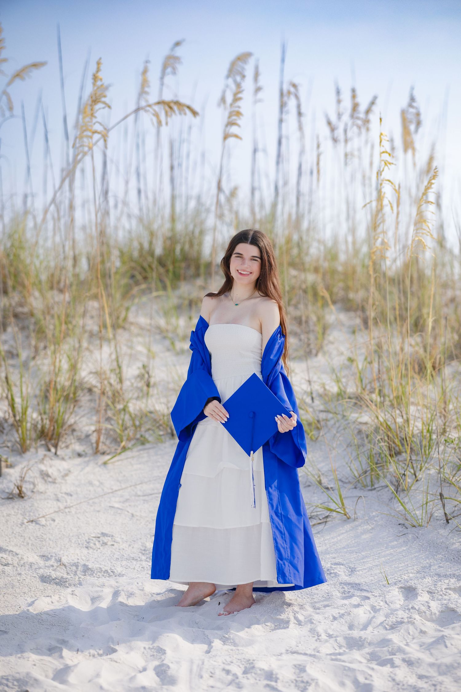 Young woman in white dress with blue graduation robe and cap on sandy beach with sea oats.