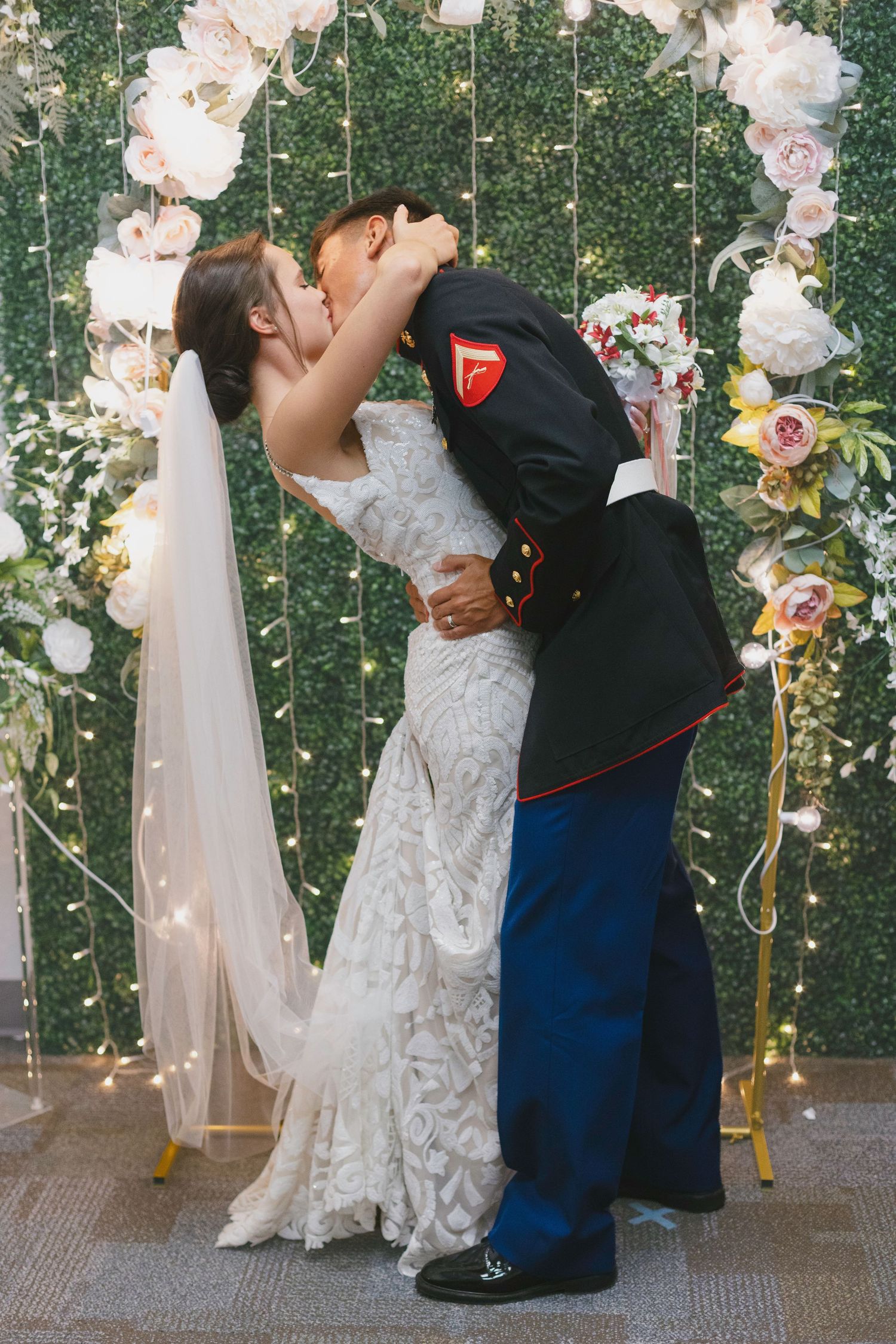 Marine groom in dress uniform kissing bride in white lace gown at floral wedding altar with fairy lights.