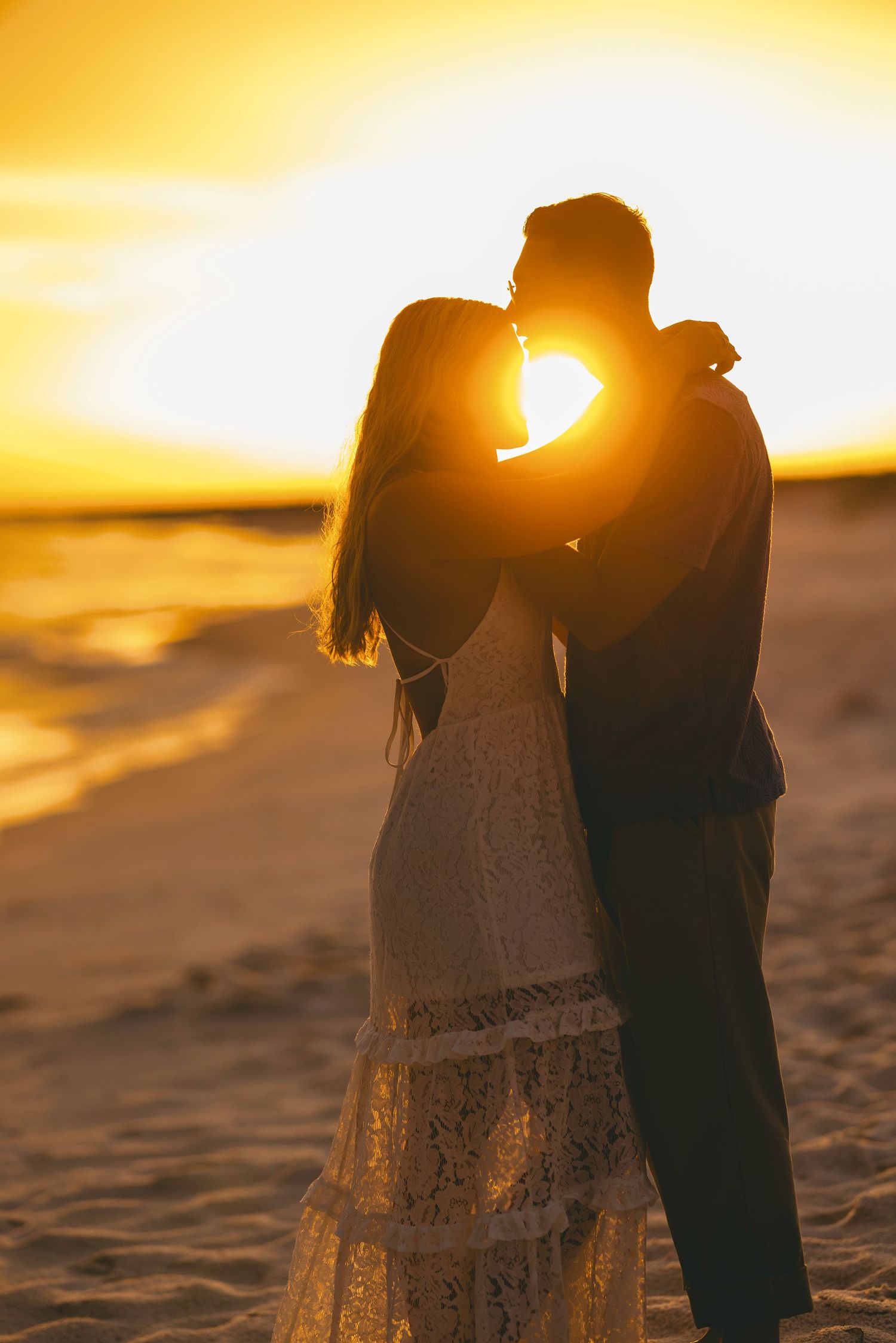 Romantic couple silhouetted against a golden sunset on the beach, bride in lace dress embracing groom.