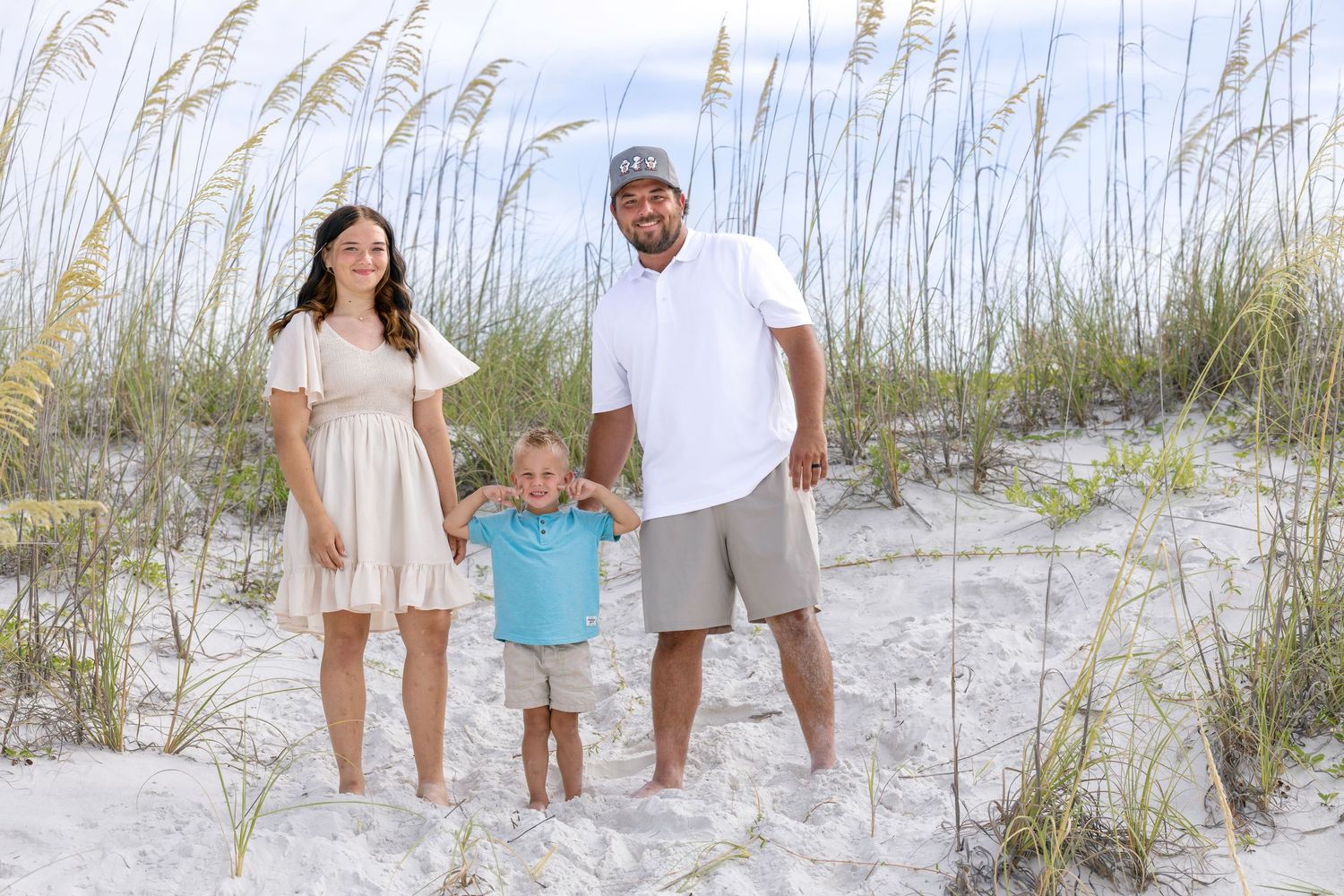 Young family of three standing among sea oats on sandy beach, toddler held between smiling parents.