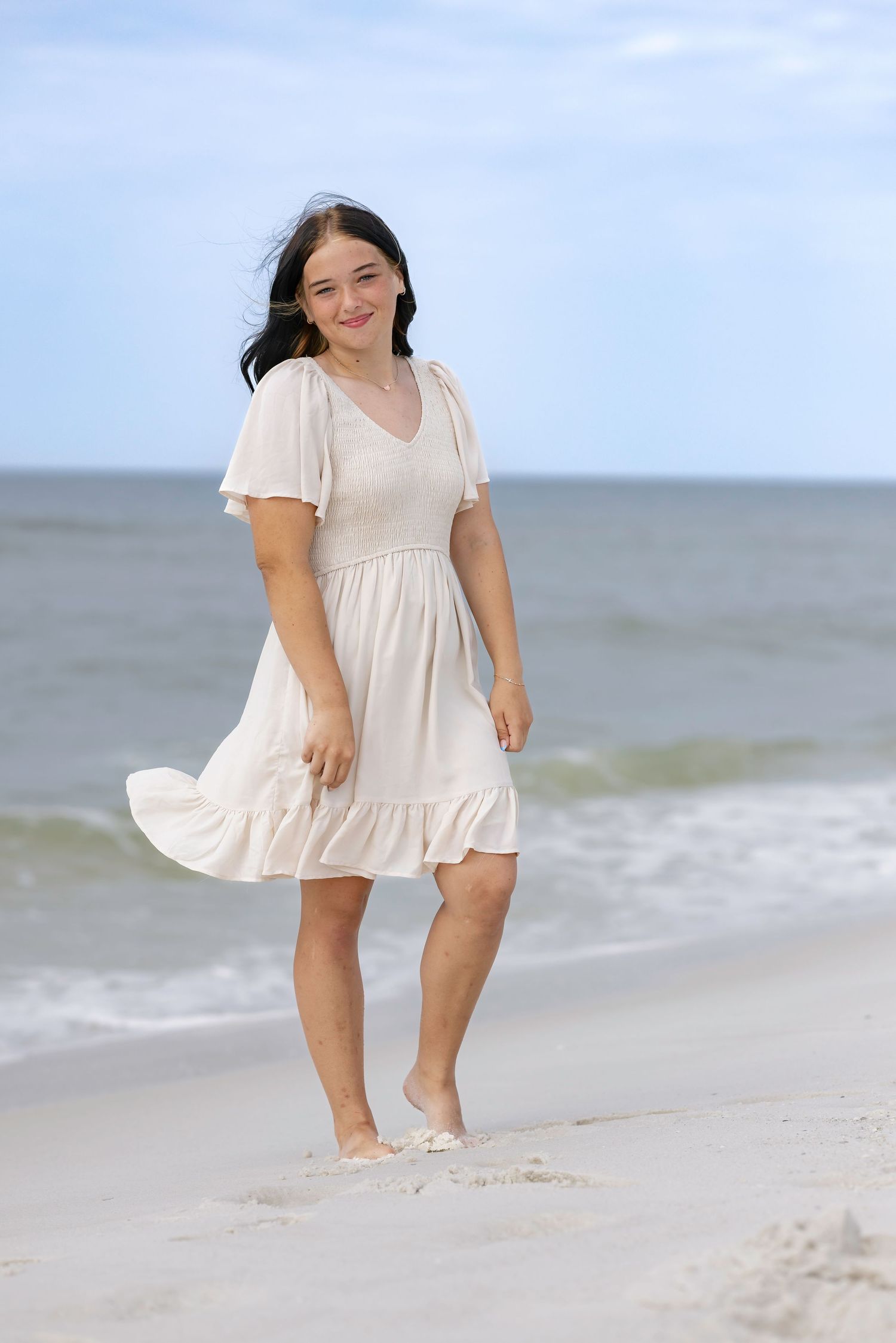 Young woman in white dress walking barefoot on sandy beach with ocean waves in background.