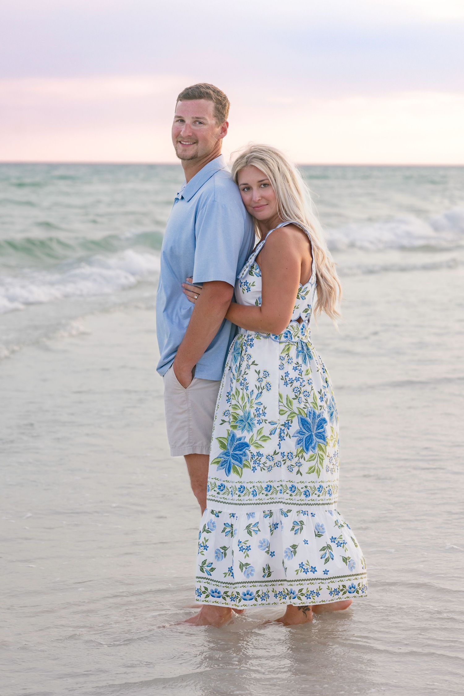 Couple embracing on a beach at sunset, woman in floral dress and man in blue shirt standing in shallow water.