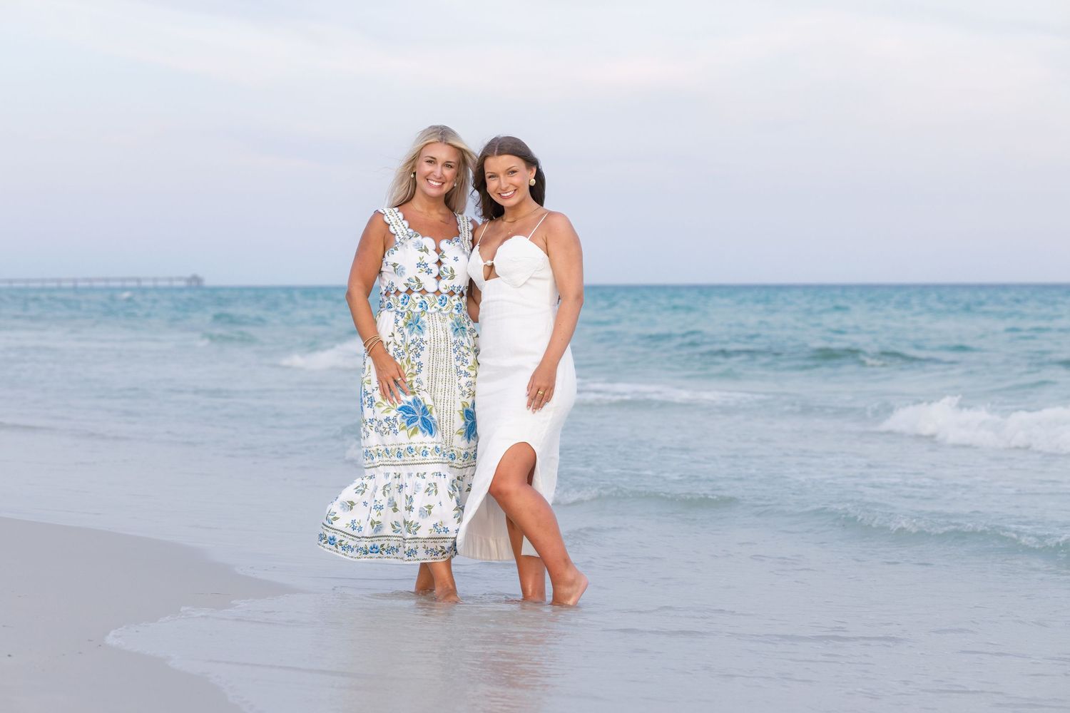 Two women in elegant dresses pose smiling on a sandy beach with ocean waves behind them.